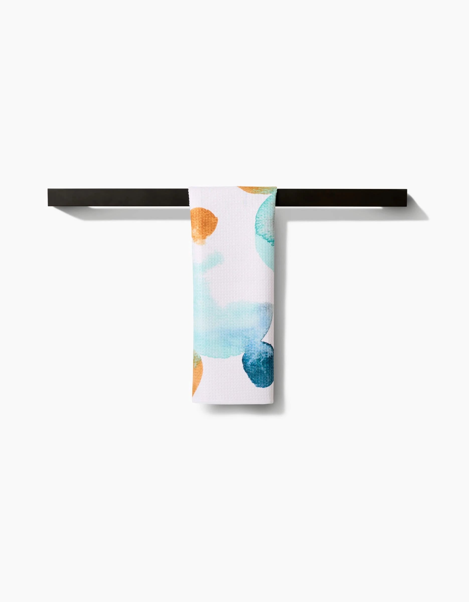The Taryn Brennan Watercolor Rocks Bar Towel, featuring blue, teal, and orange spots on white and made from recycled materials, hangs neatly over a black bar against a plain white background.