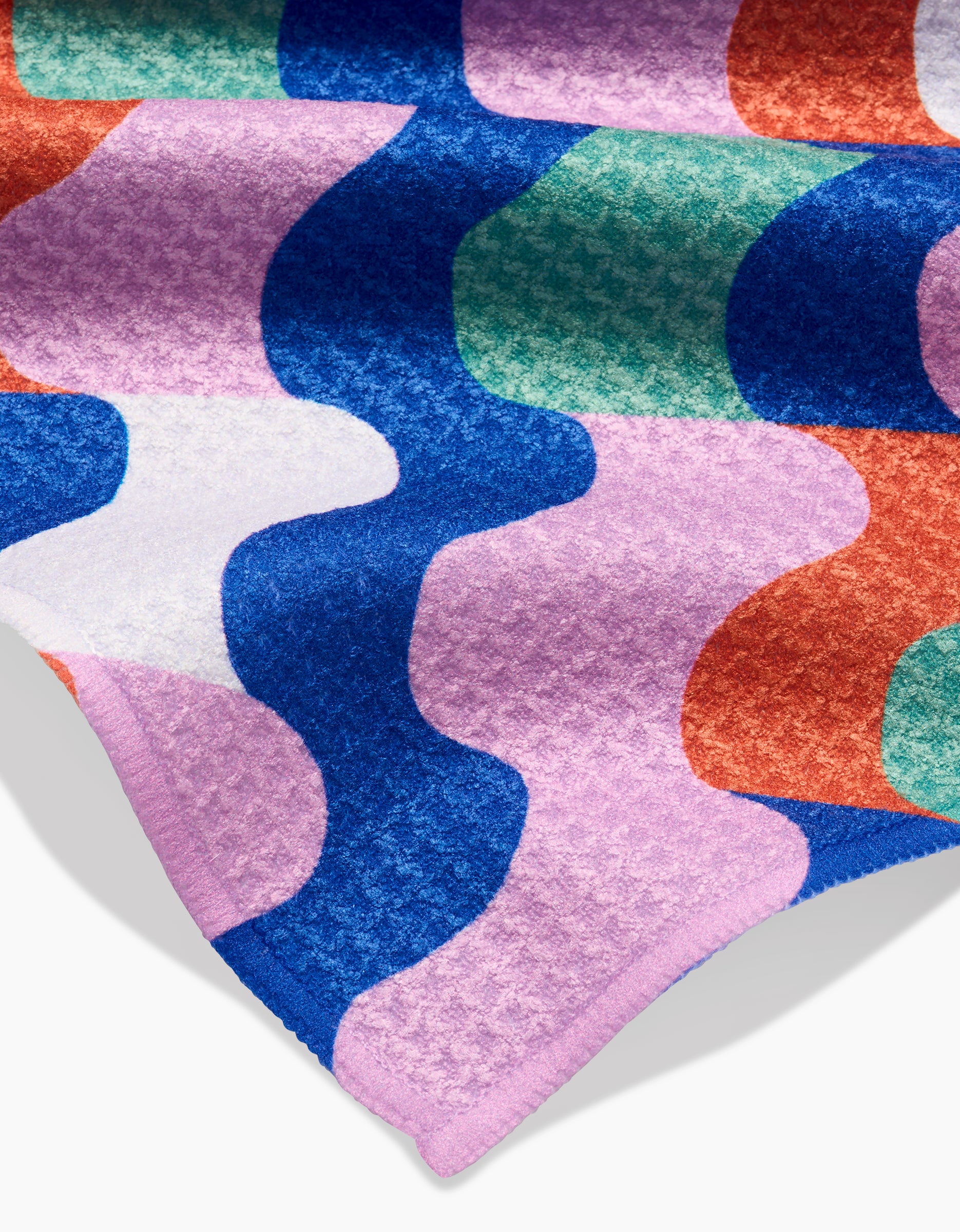 A close-up of the “Wavy Stripes” bar towel by Claire Iglesias, featuring colorful, overlapping Geoweave patterns in blue, red, pink, green, and white. A corner—made from post-consumer recycled material—is folded over.