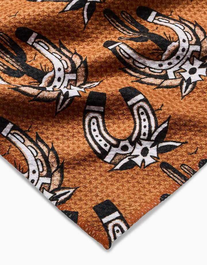 A close-up of Cole Strems Western Horseshoes bar towel, featuring textured brown Geoweave fabric with a black and white southwestern pattern of horseshoes, stars, and cacti.