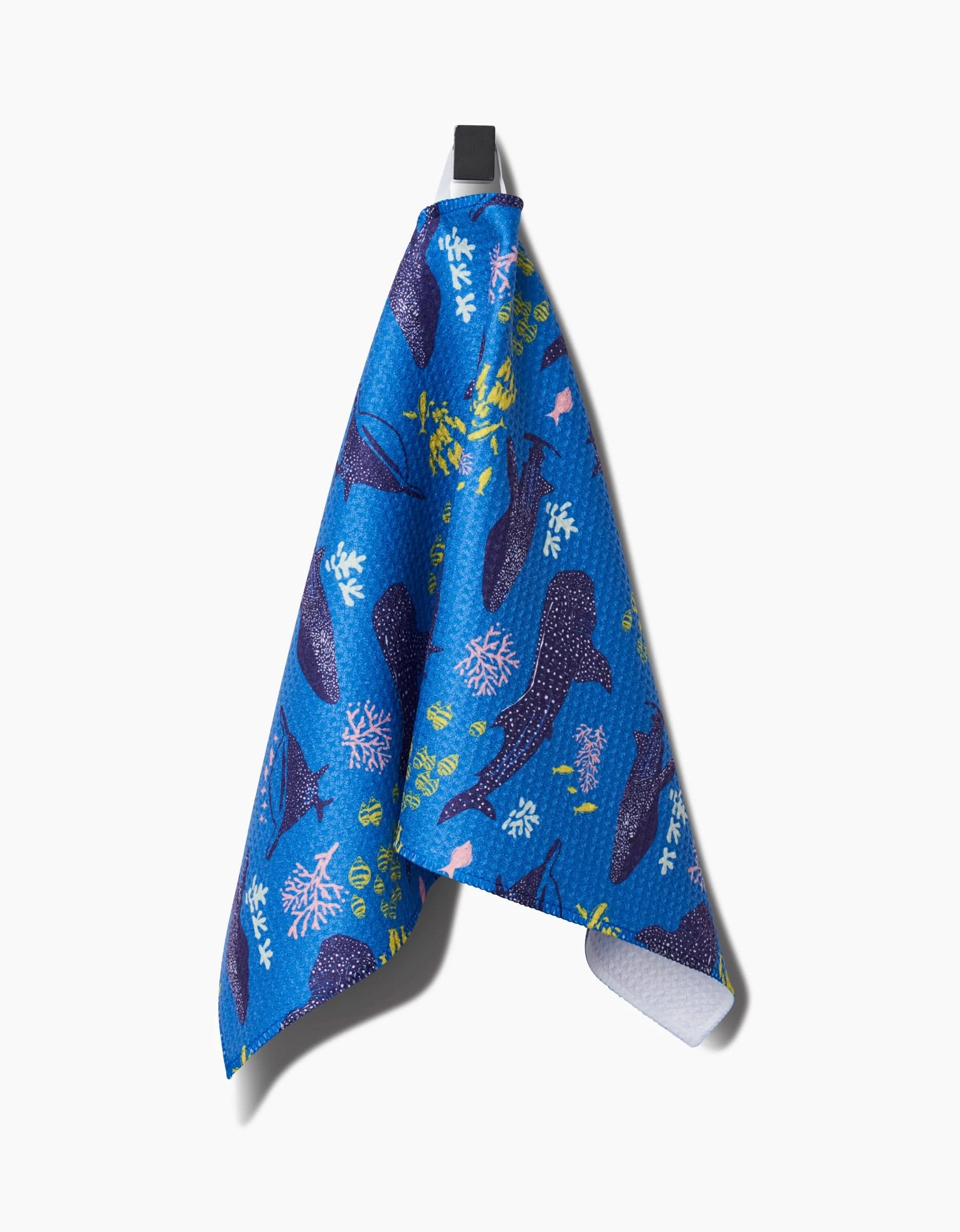 A Whale Shark Bar Towel by Elisabeth Wing, featuring vibrant ocean-inspired patterns on Geoweave fabric, hangs from a black hook against a plain white background.