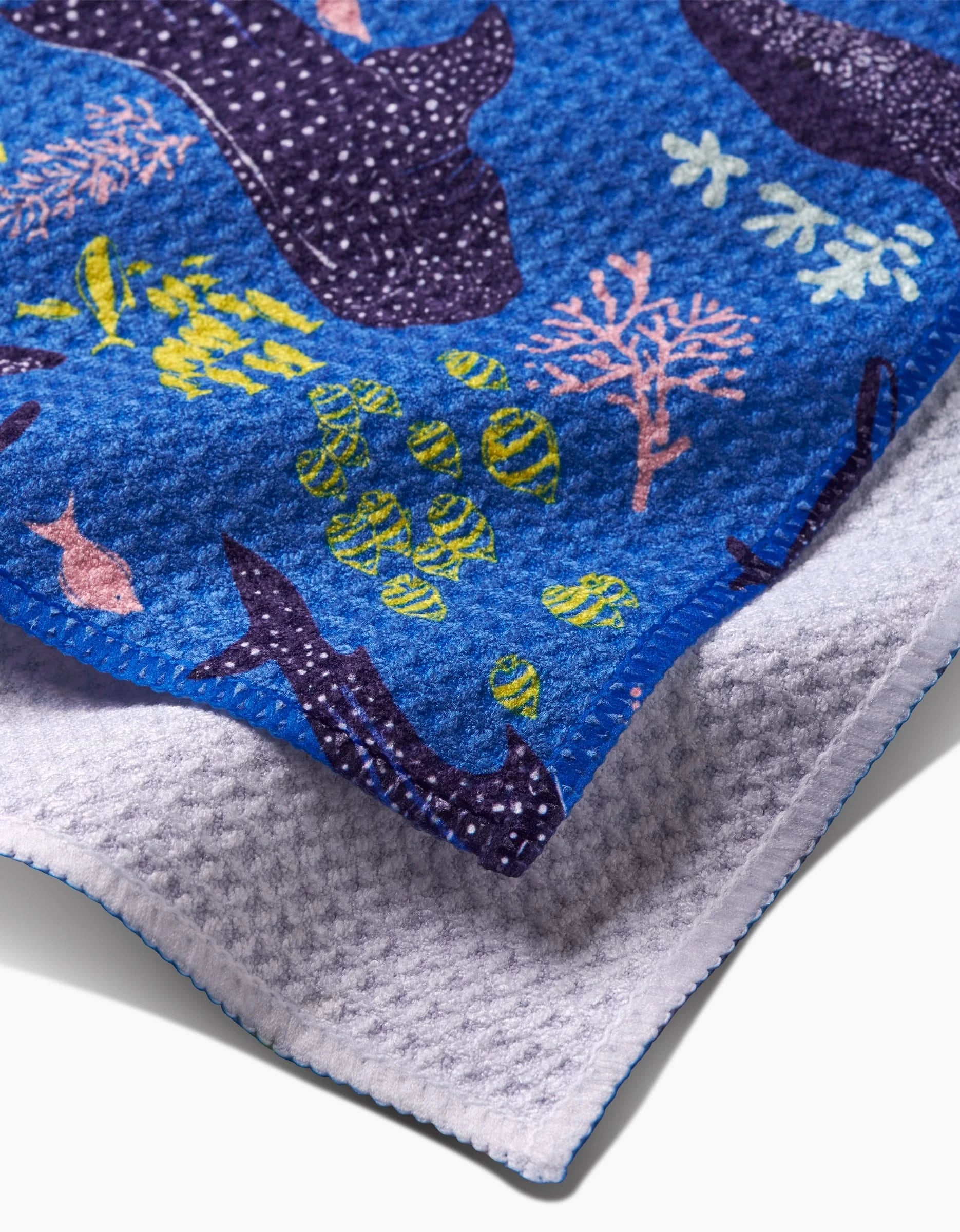 A close-up of the Elisabeth Wing Whale Shark towel, textured and made from post-consumer recycled materials, showcases a vibrant underwater scene with fish, coral, and whales, layered over a plain white towel.