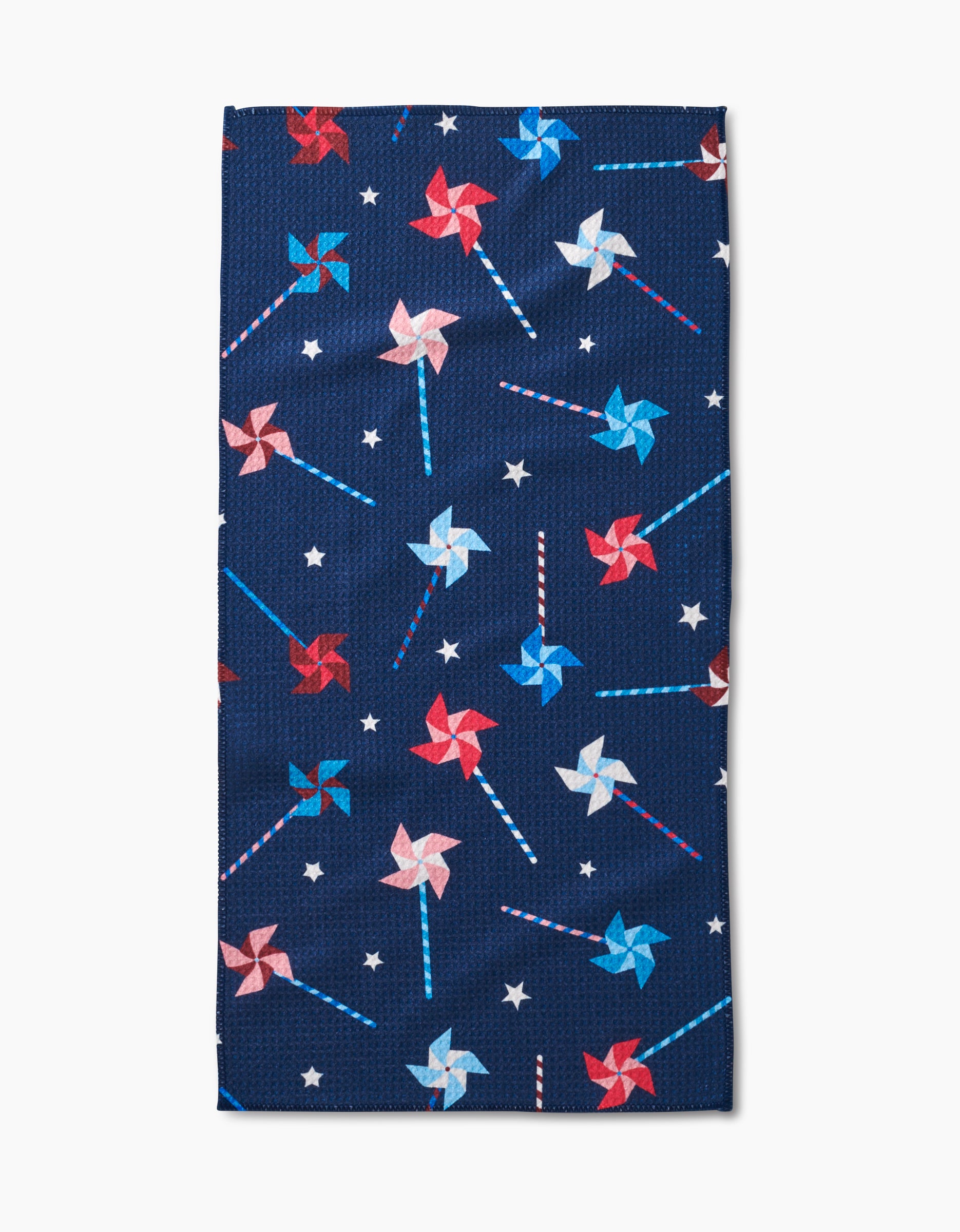 The Whirlwind Pins bar towel by Rebecca Bobko is navy blue, made from recycled materials, and features a festive pattern of red, white, and blue pinwheels with small white stars.