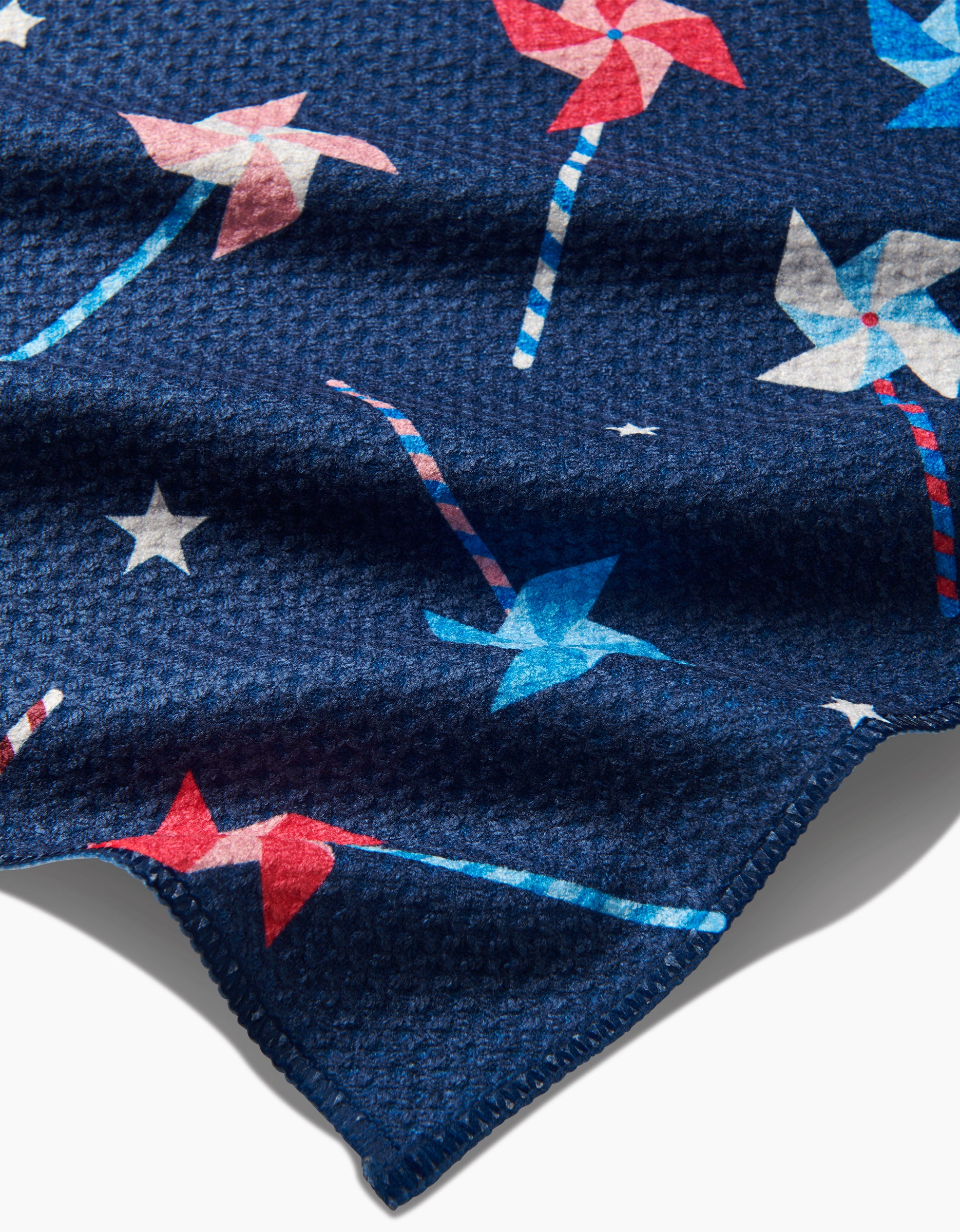 A close-up of Rebecca Bobko’s Whirlwind Pins Bar Towel shows dark blue Geoweave fabric with red, white, and blue pinwheels, white stars, and a textured waffled surface. The towel’s edge appears in the foreground.