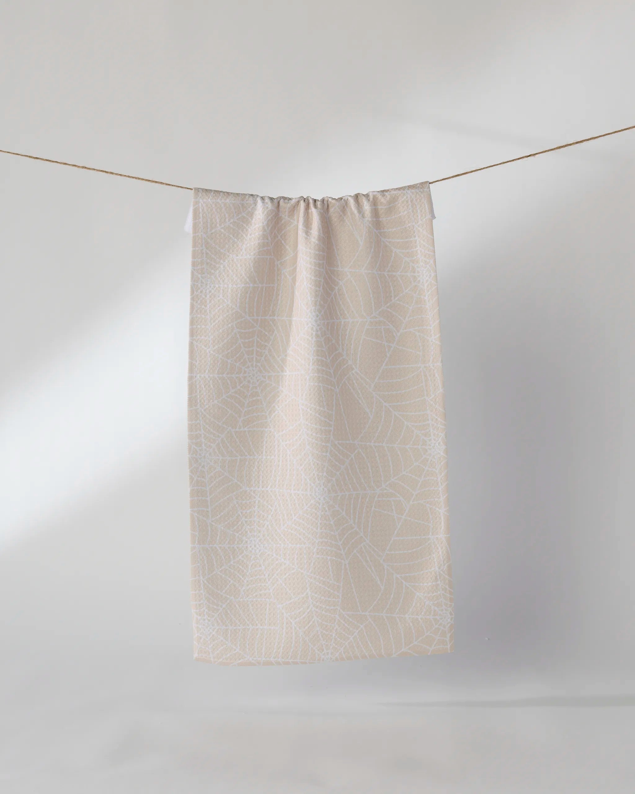 The Geometry Whisper Web towel, in beige with a delicate white spiderweb design, hangs from a string against a simple, light background.