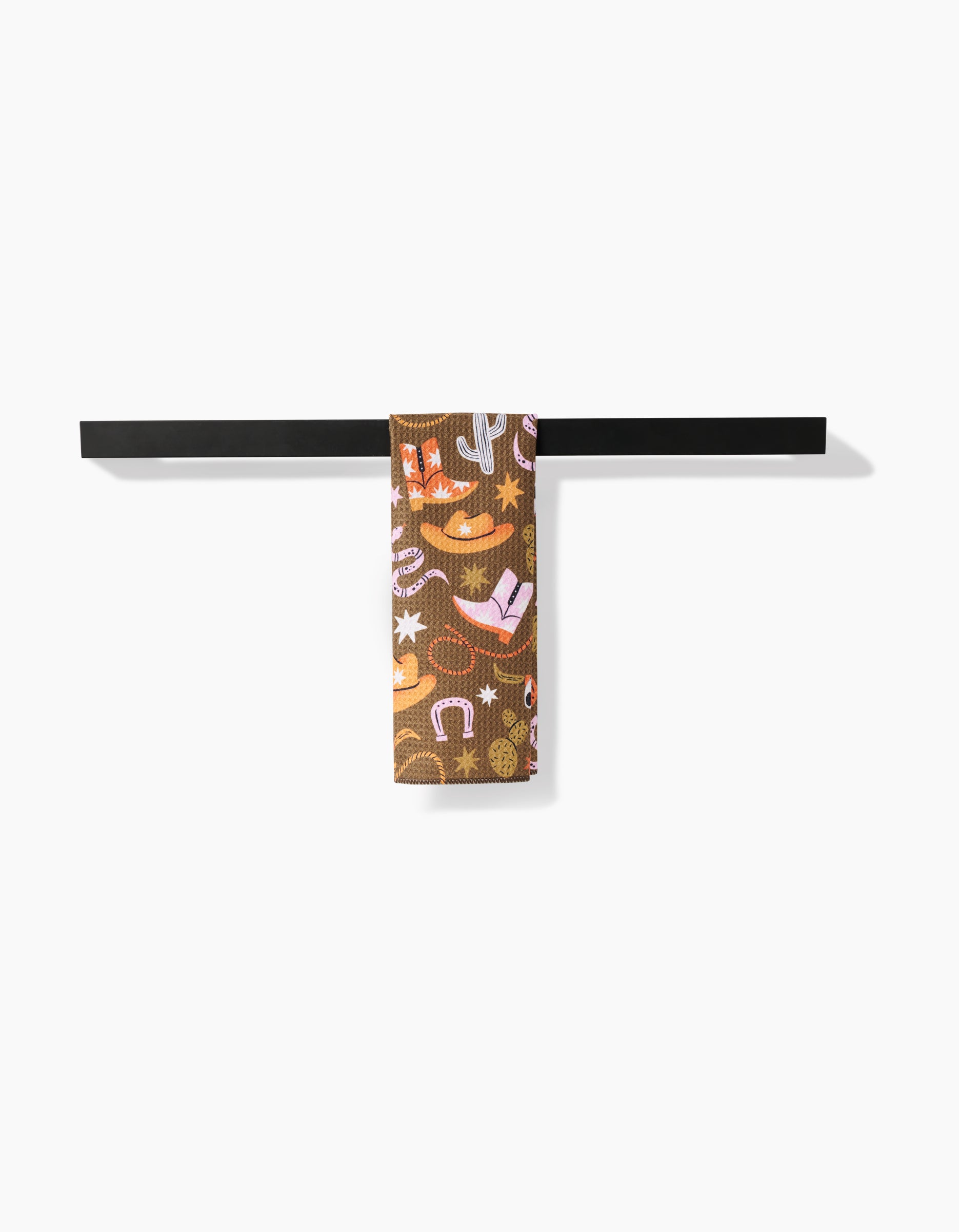 The Megan Roy Wild Cowboy bar towel, featuring a vibrant cowboy-themed design and made from recycled materials, hangs on a sleek black towel bar against a white background.