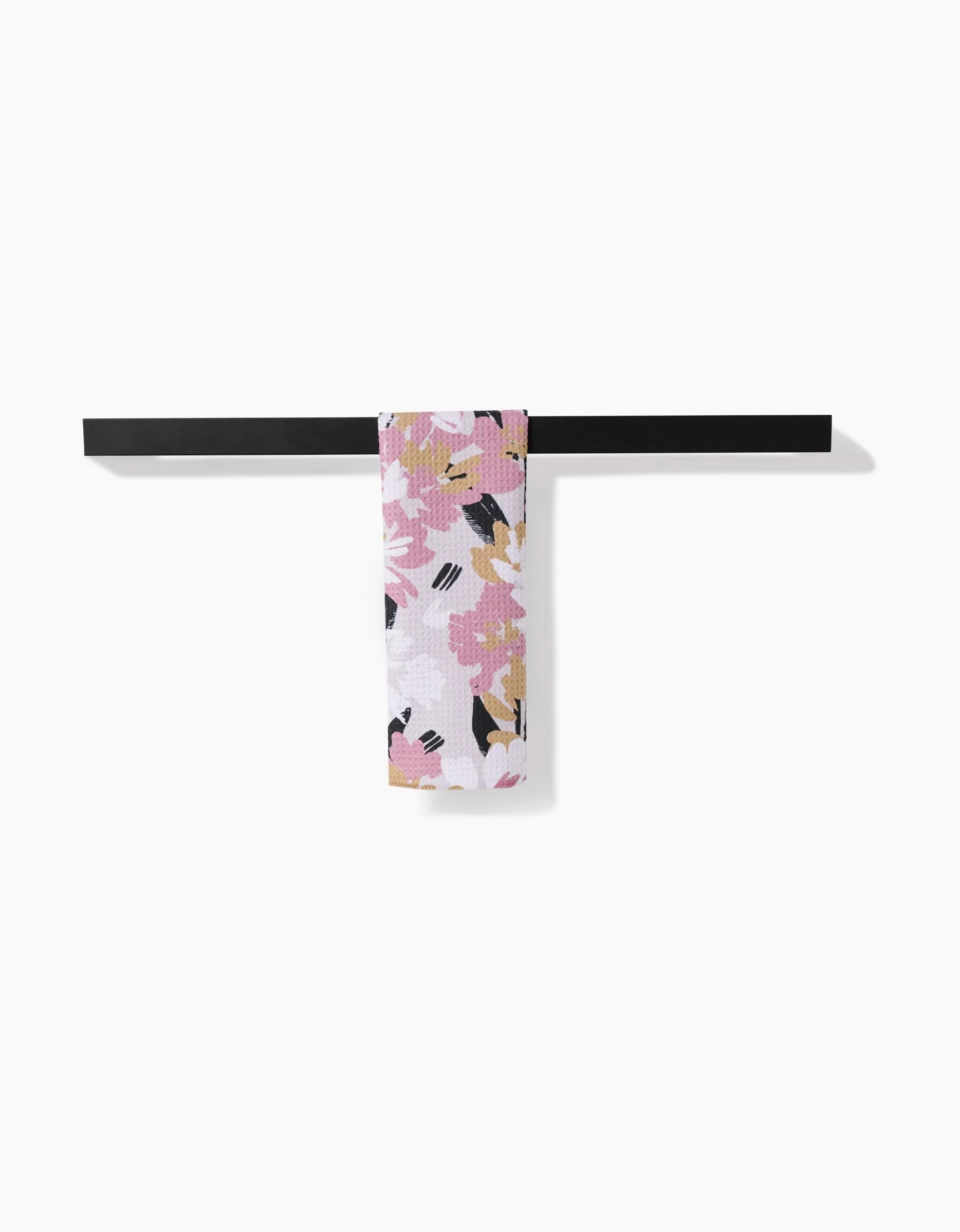 The Toboggan Avenue Wilderness Floral Bar Towel, made from recycled materials in pink, white, black, and mustard florals, is neatly draped over a minimalist black towel bar against a plain white background.