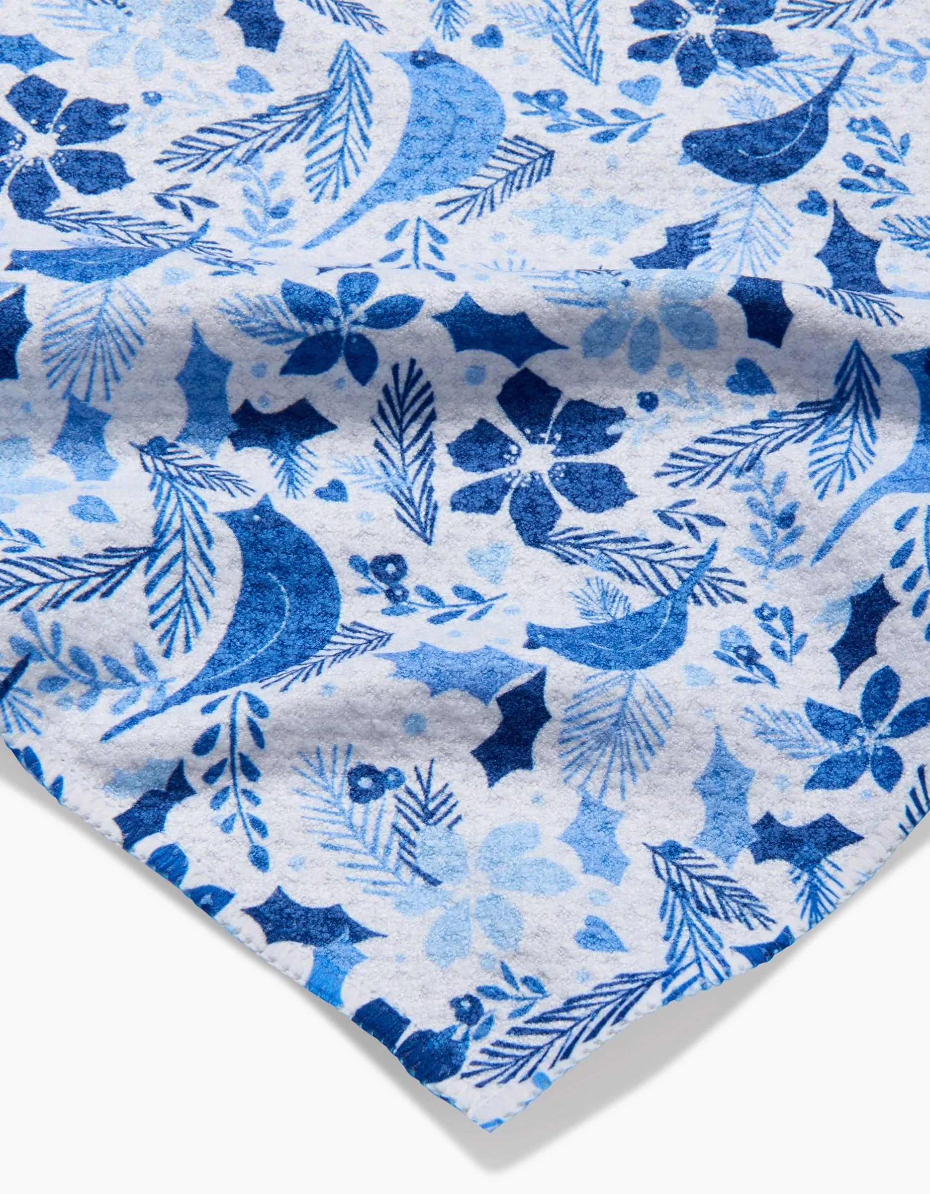 A close-up of Kris Ruff’s Winter Blue Birds fabric shows its blue and light blue floral and leaf pattern on white; the texture and folded corner highlight its appeal for ultra-absorbent towels made from recycled materials.