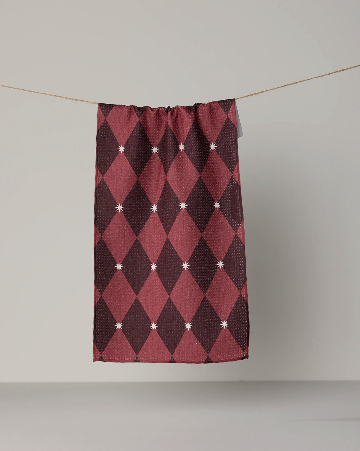 The Geometry “Winter Gems” mini towel, featuring a maroon diamond pattern adorned with small white star accents, hangs on a clothesline against a plain light gray background. Made from premium Geoweave® fabric, this towel combines style with lasting durability.
