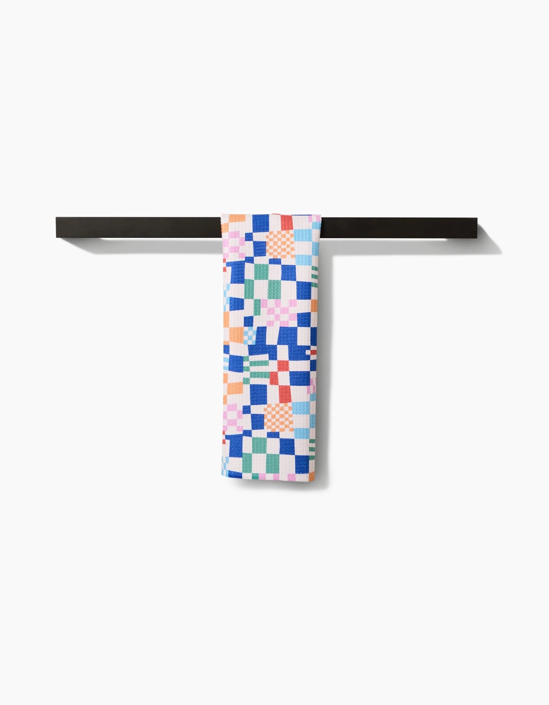 The Wonky Checkers bar towel by Claire Iglesias, featuring a colorful patchwork pattern on Geoweave fabric, hangs neatly over a minimalist black bar against a plain white background.