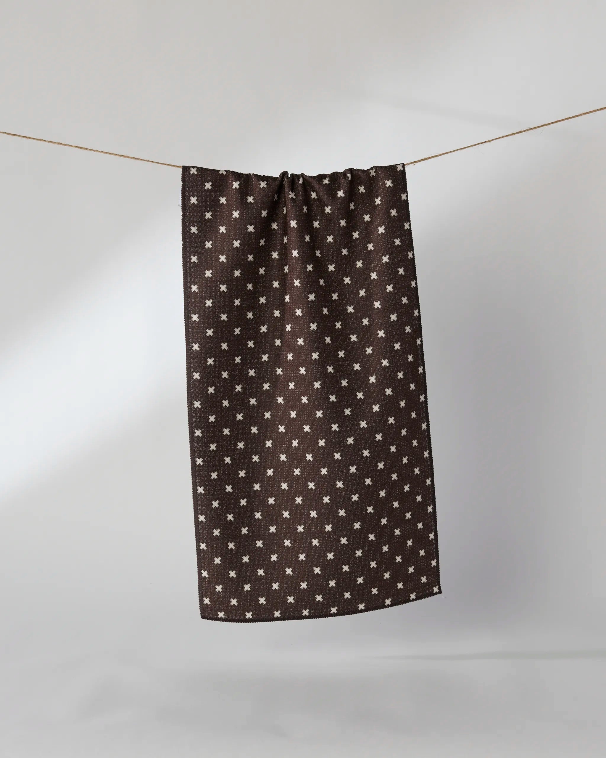 The Geometry Woven Spice bar towel, dark brown with a white cross pattern and made from recycled materials, hangs from a string against a white background.