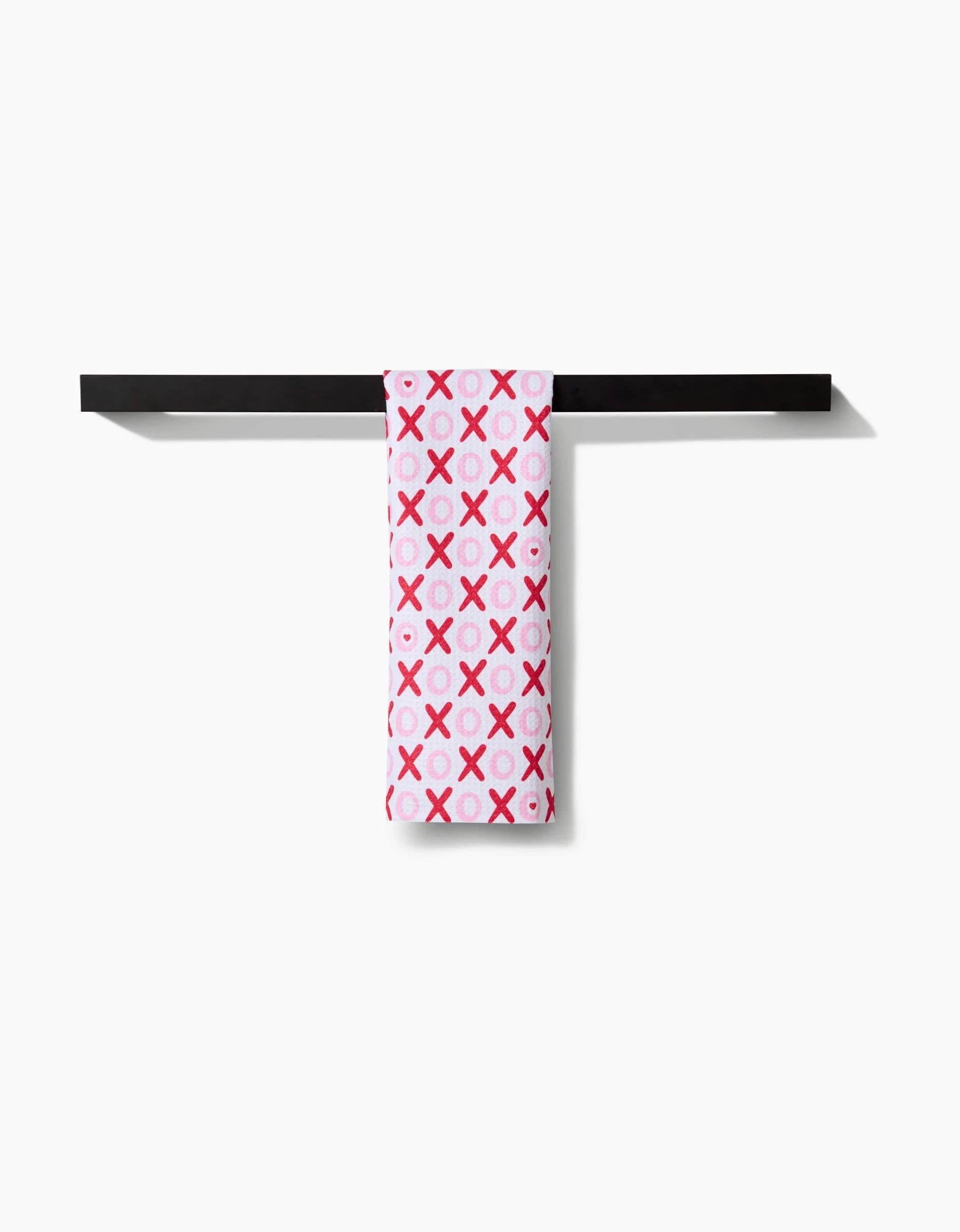 The XOXO bar towel by Sarah Daugherty features a pink fabric with a red X and O pattern, draped neatly over a black horizontal towel bar against a white background.