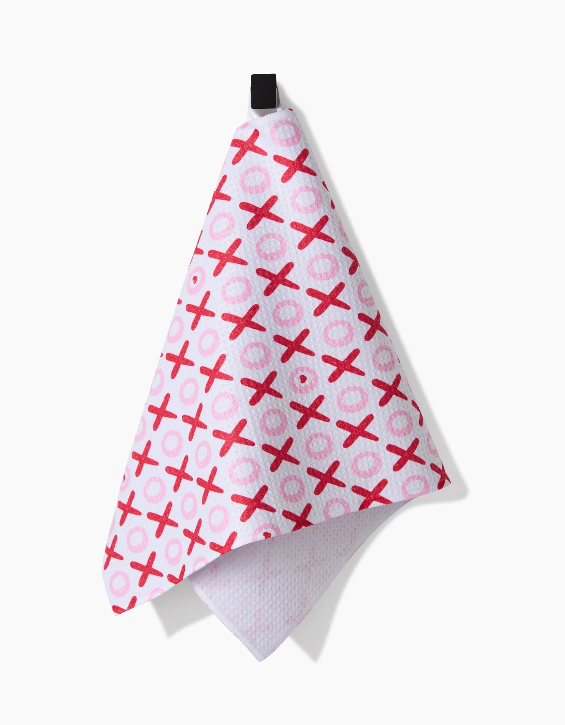The XOXO towel by Sarah Daugherty, featuring a red and pink X and O pattern, hangs from a black loop against a plain white background.