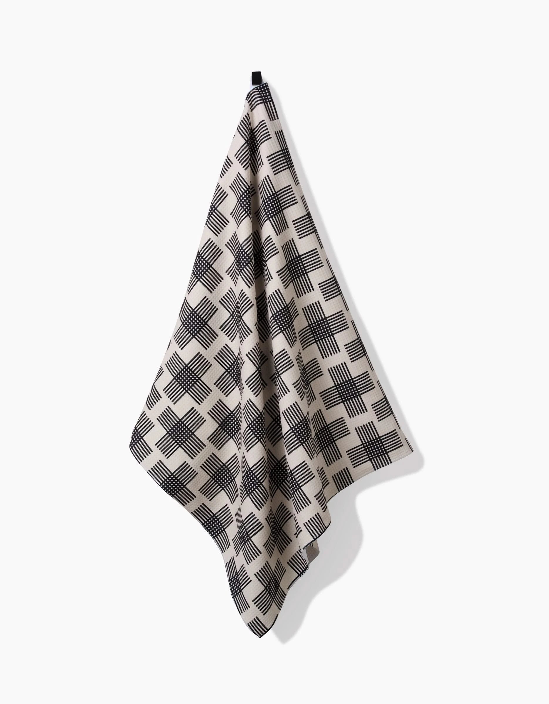 The Cross Hatch bath towel by Rosa Kichline, in beige with a black crisscross design and ultra-absorbent Geoweave technology, hangs from a small hook against a plain white background.