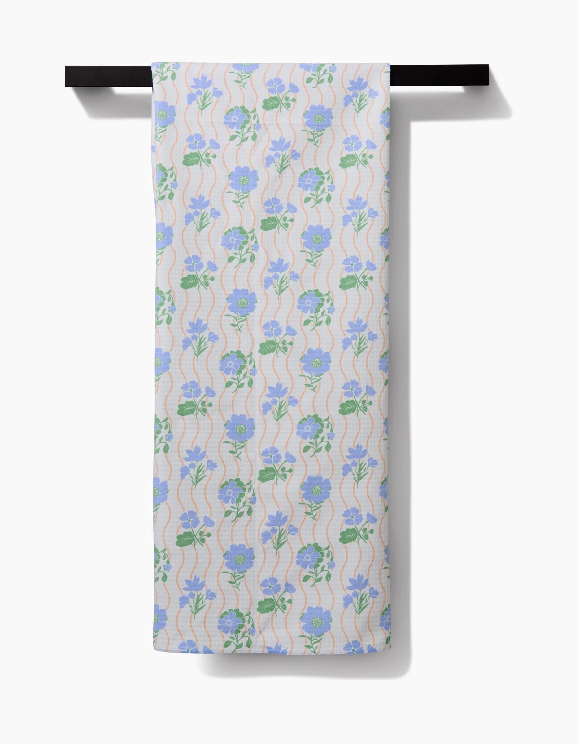 The Danelys Sidron Spring Wave Luxe Bath Towel hangs on a black rod, displaying blue and purple florals with green leaves and peach waves, all on a light background. Made from ultra-absorbent post-consumer recycled materials.