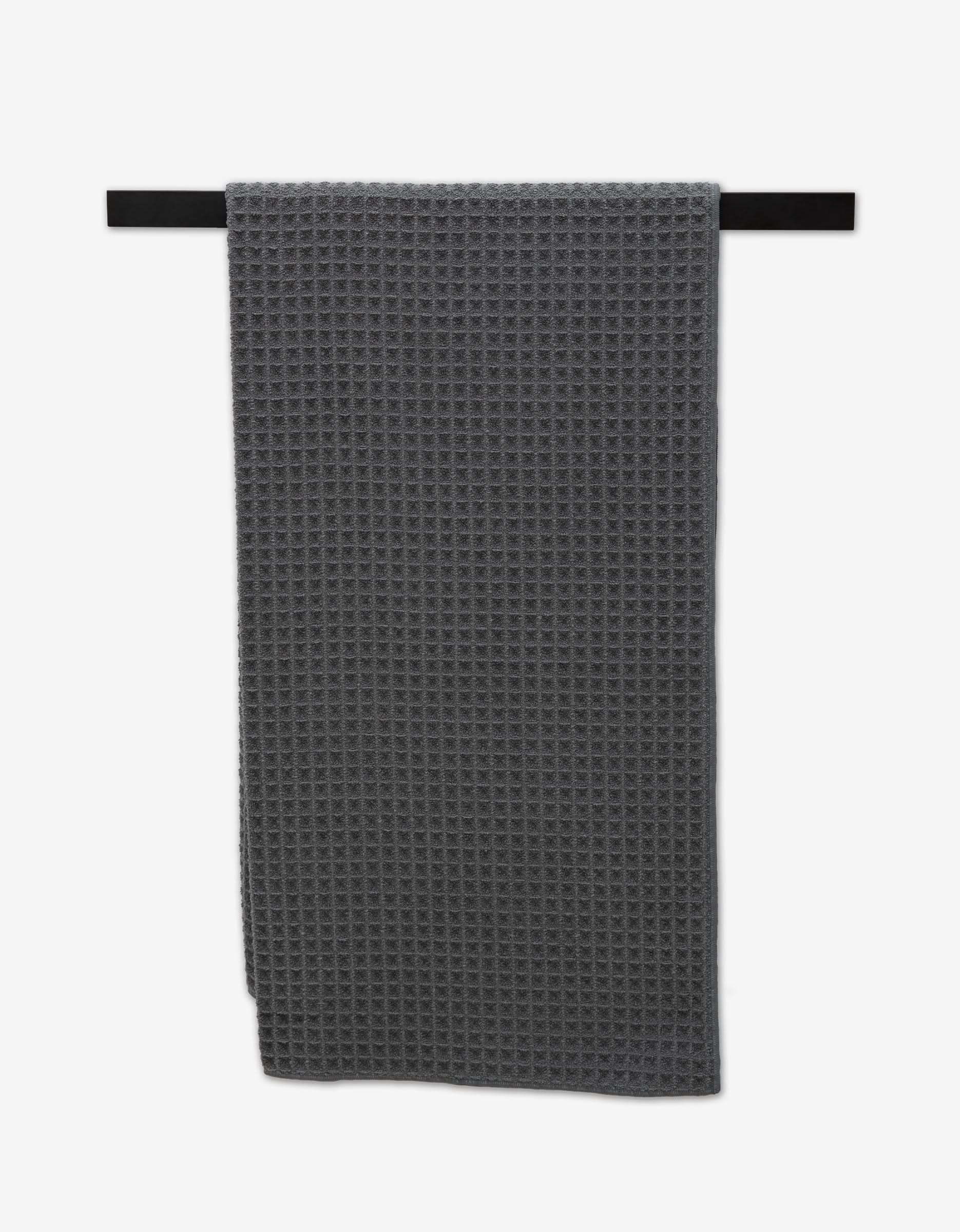 The GEOMETRY Charcoal Waffle Bath Towel, made from post-consumer recycled materials, hangs neatly on a black towel bar against a white background.