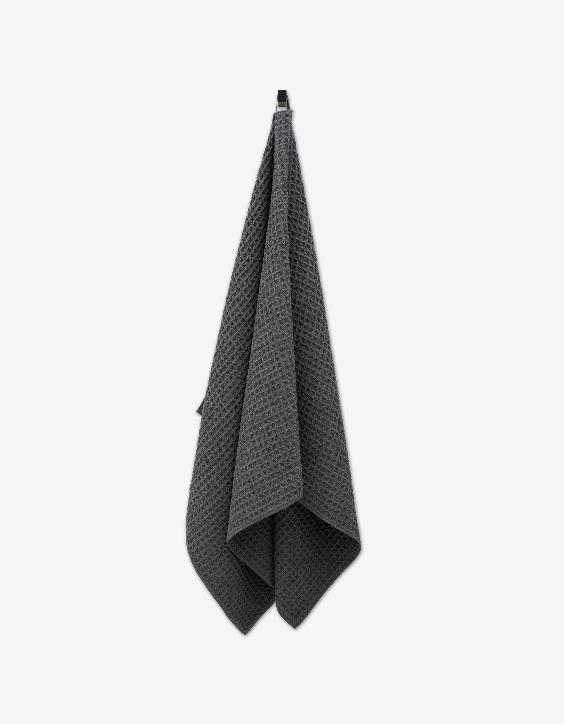A Charcoal Waffle Bath Towel by GEOMETRY, made from post-consumer recycled materials, hangs from a black hook against a plain white background.