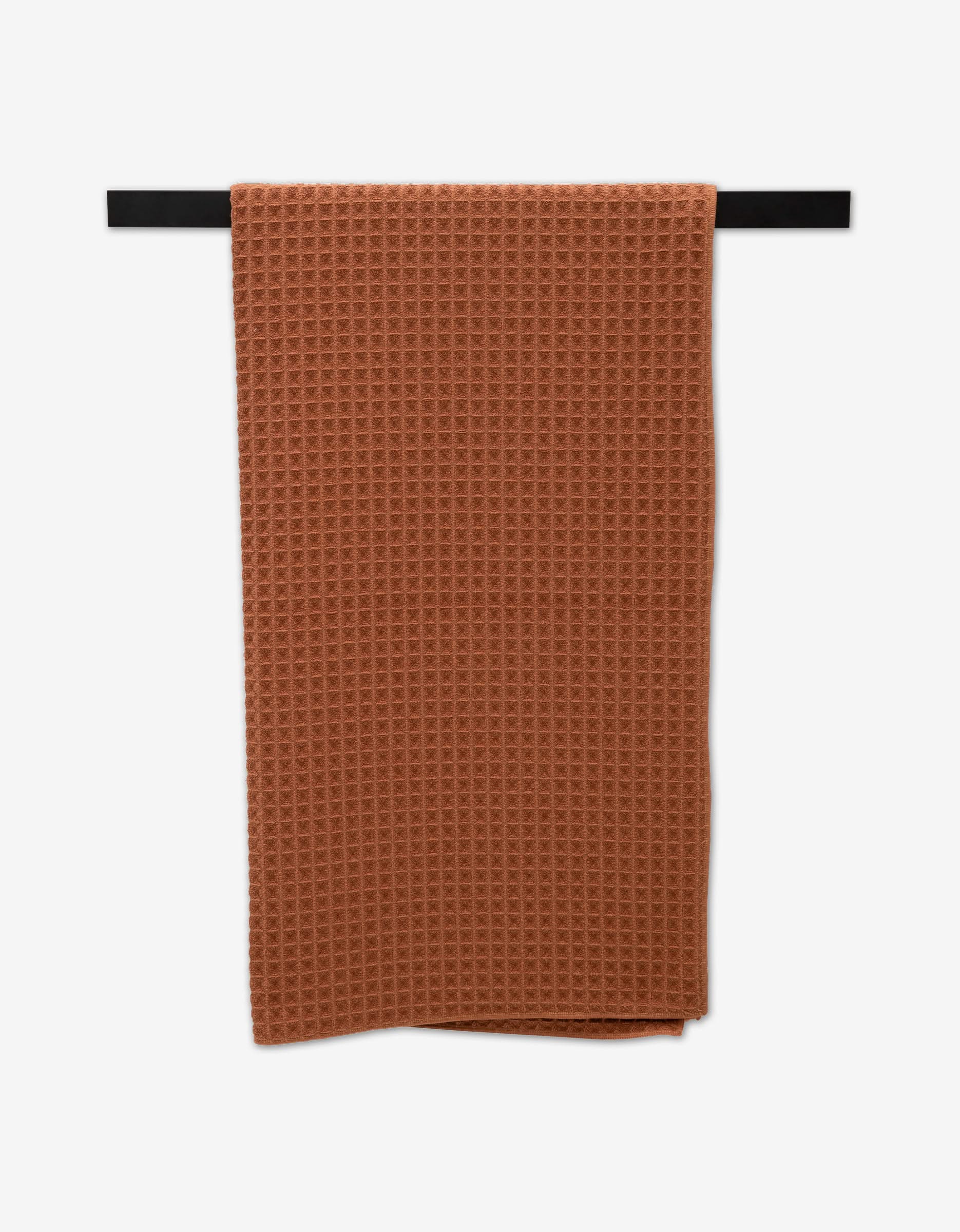 A Clay ultra-absorbent Waffle Bath Towel by GEOMETRY, in a rich rust-orange hue, is neatly folded and hanging on a black horizontal bar against a white background.