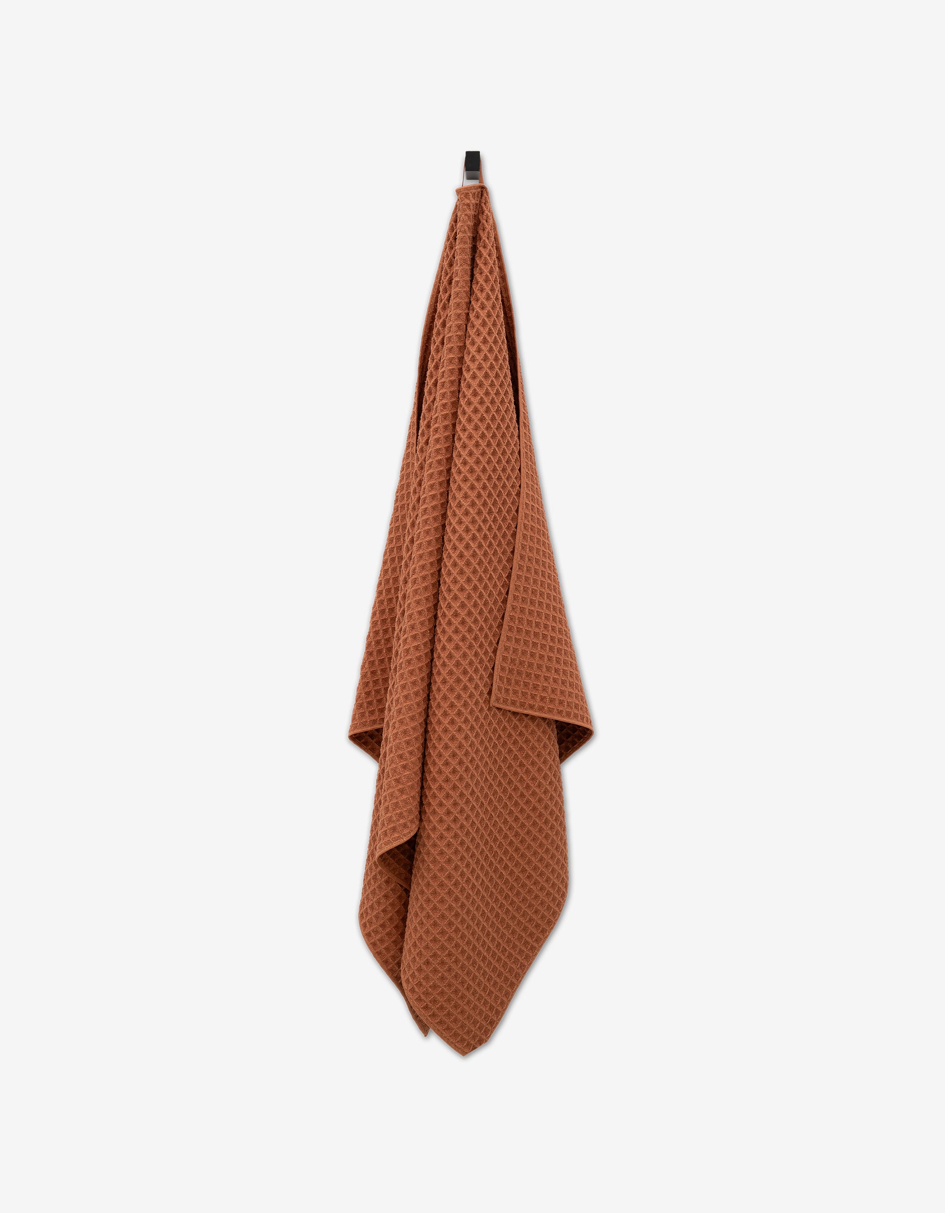The GEOMETRY Clay Waffle Bath Towel, featuring a luxe waffle weave in a rust hue, hangs from a hook against a white background, highlighting its ultra-absorbent quality.
