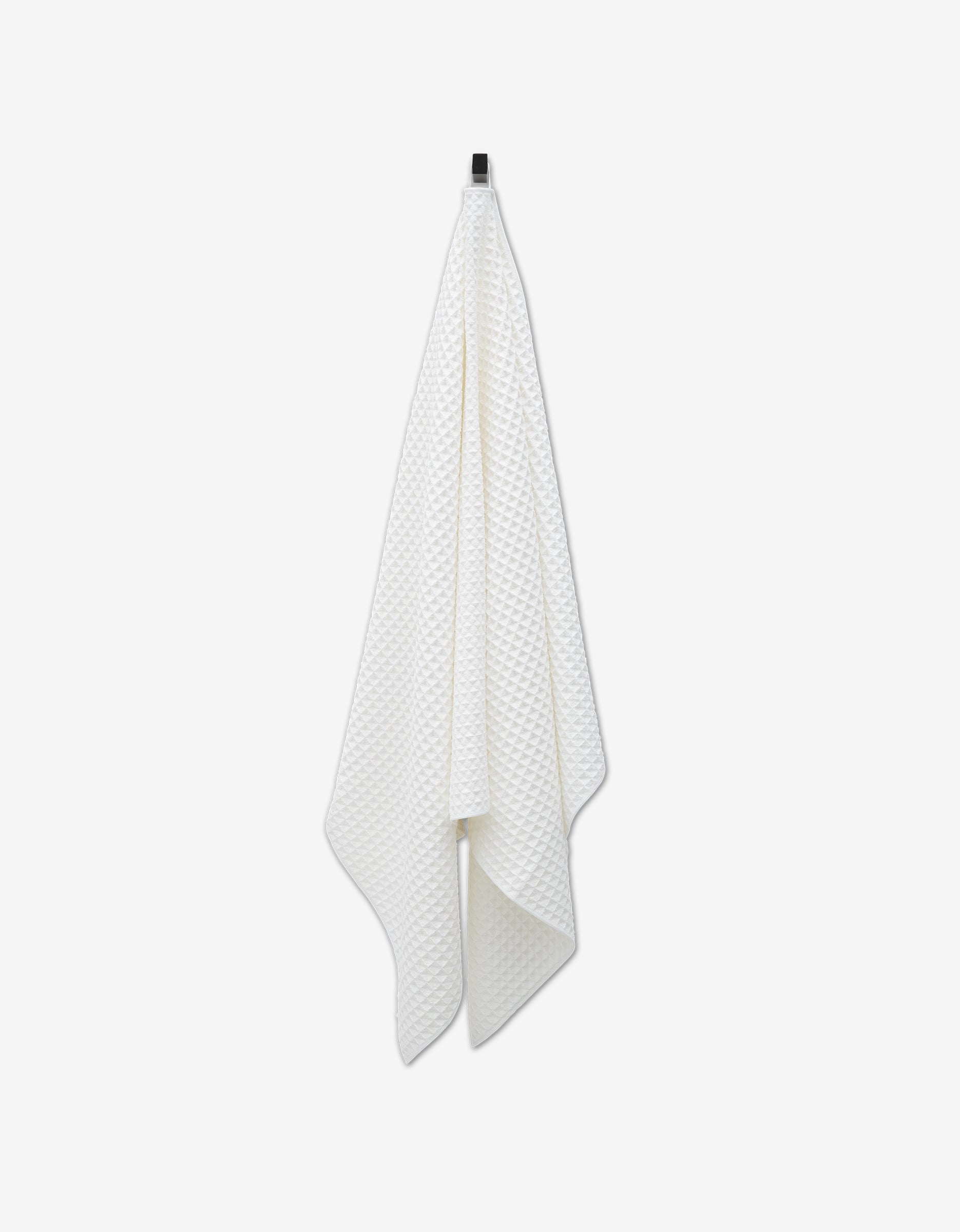 The GEOMETRY Cloud towel, a white textured waffle bath towel crafted from recycled materials, hangs on a black hook against a plain white background.