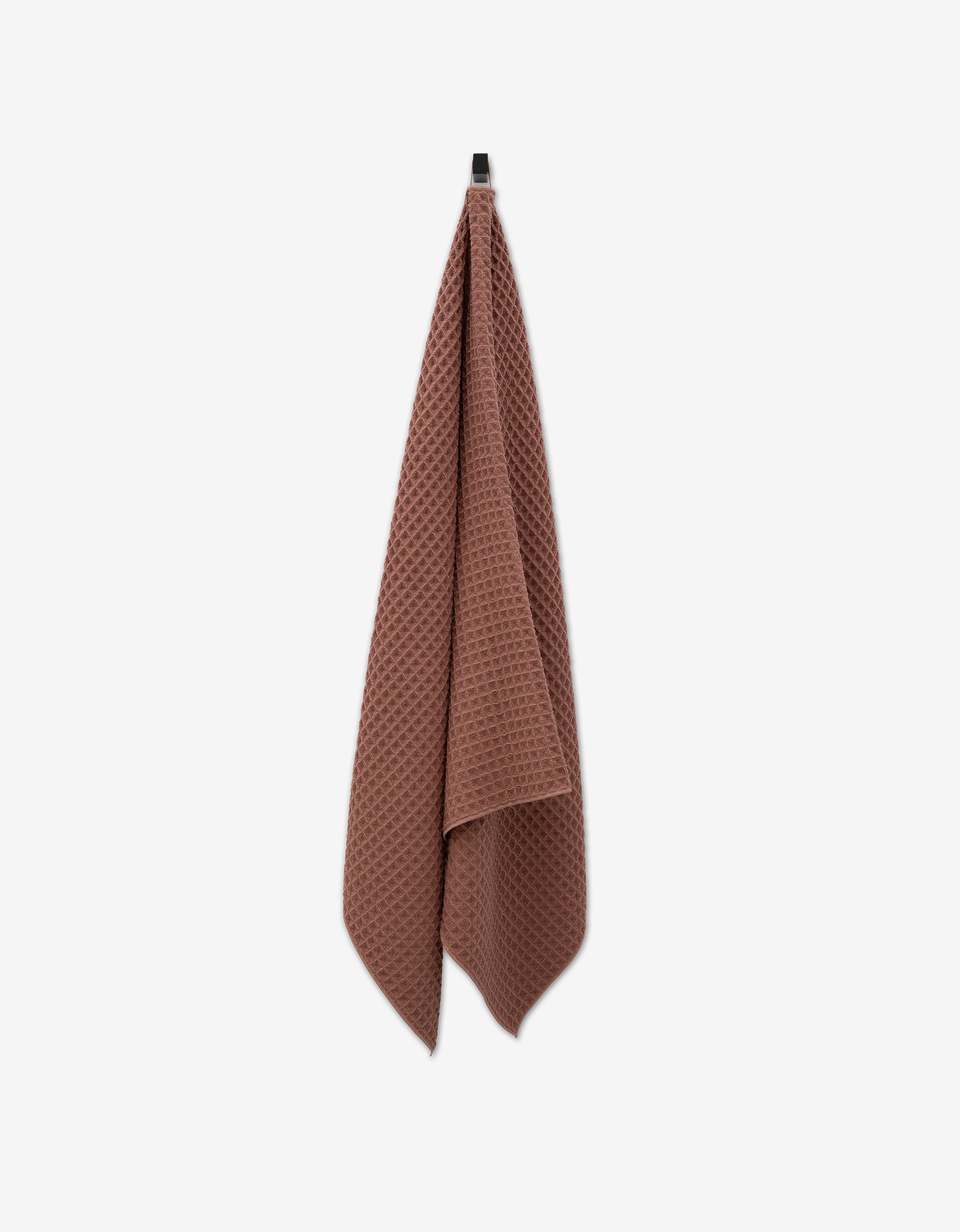 A GEOMETRY Dusty Rose waffle bath towel hangs from a black hook against a plain white background, highlighting the luxury towel’s ultra-absorbent texture.