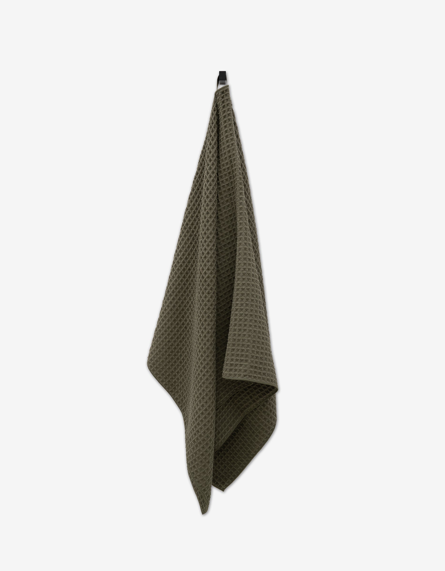 The GEOMETRY Forest towel, featuring a dark green waffle pattern, hangs on a hook against a plain white background—an eco-friendly choice that blends sustainability with classic style.