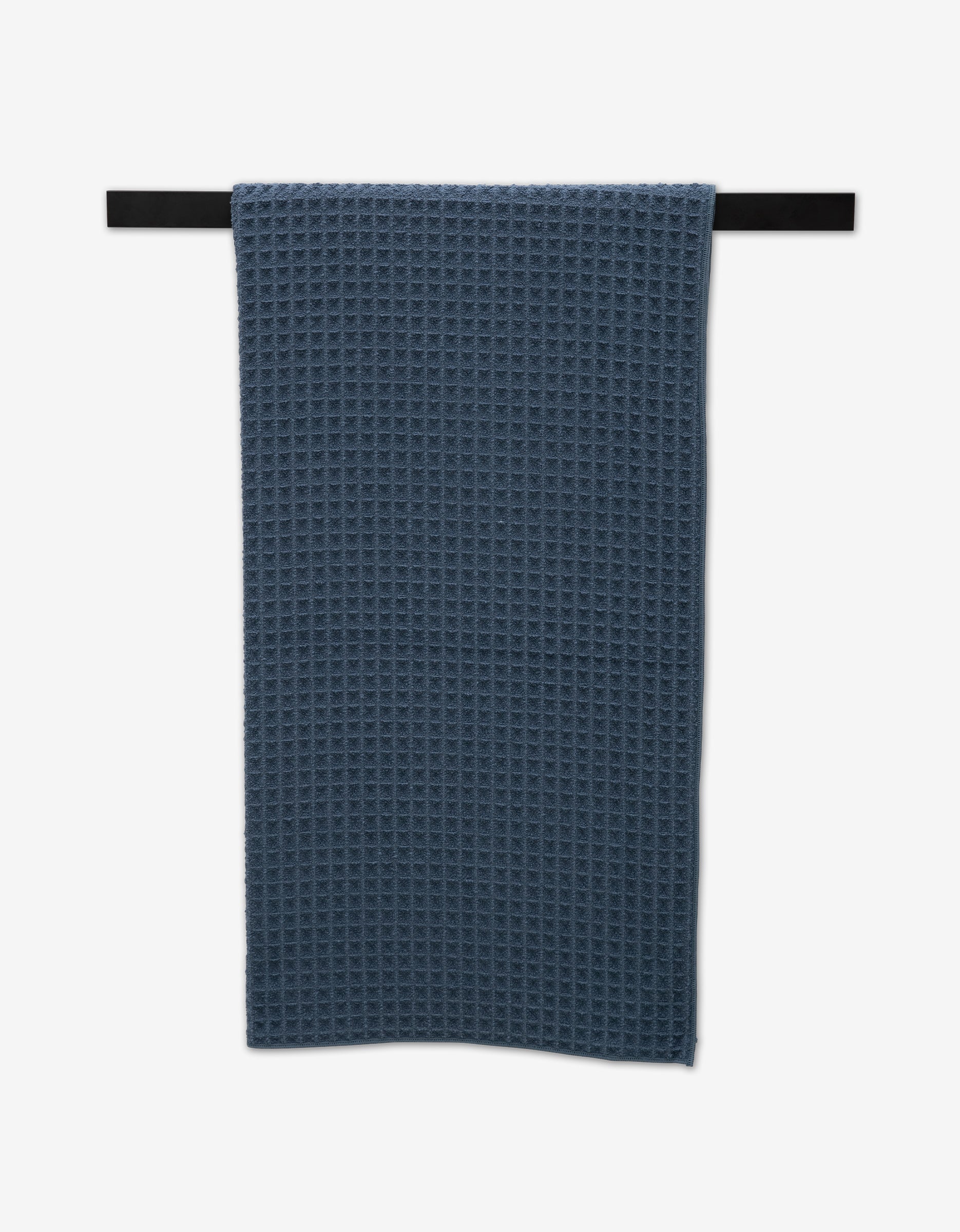 A GEOMETRY Midnight Blue Waffle Bath Towel hangs neatly on a horizontal black towel bar against a plain white background, highlighting the elegance of luxury bath towels.