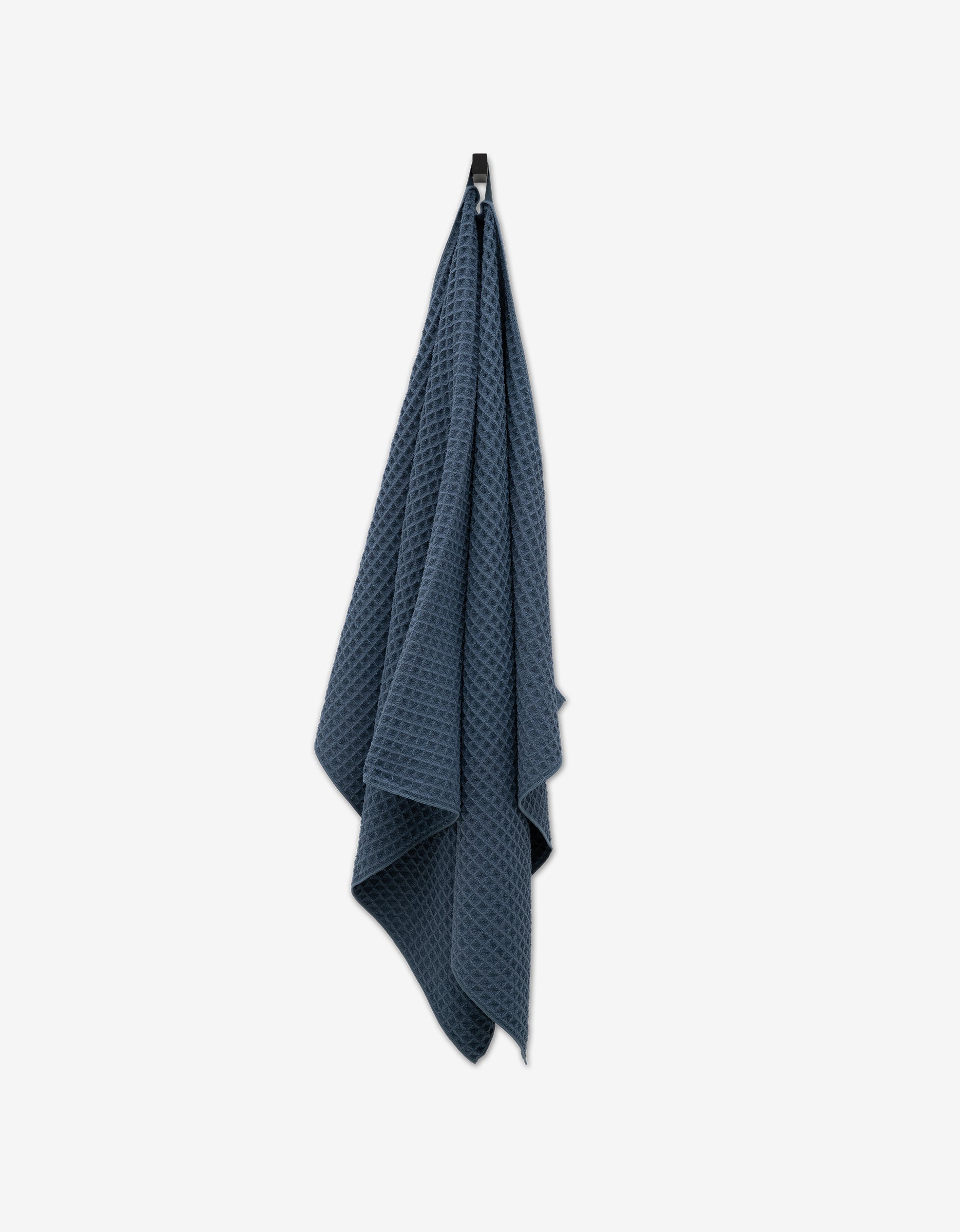 A Midnight Blue Waffle Bath Towel by GEOMETRY with a luxurious texture hangs on a black hook against a plain white background.