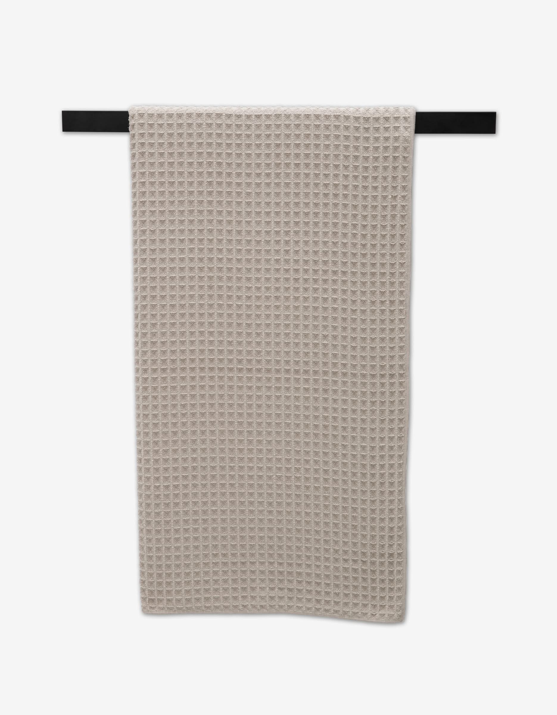 A taupe waffle-textured GEOMETRY Luxury Towel hangs neatly on a black towel bar against a white background.
