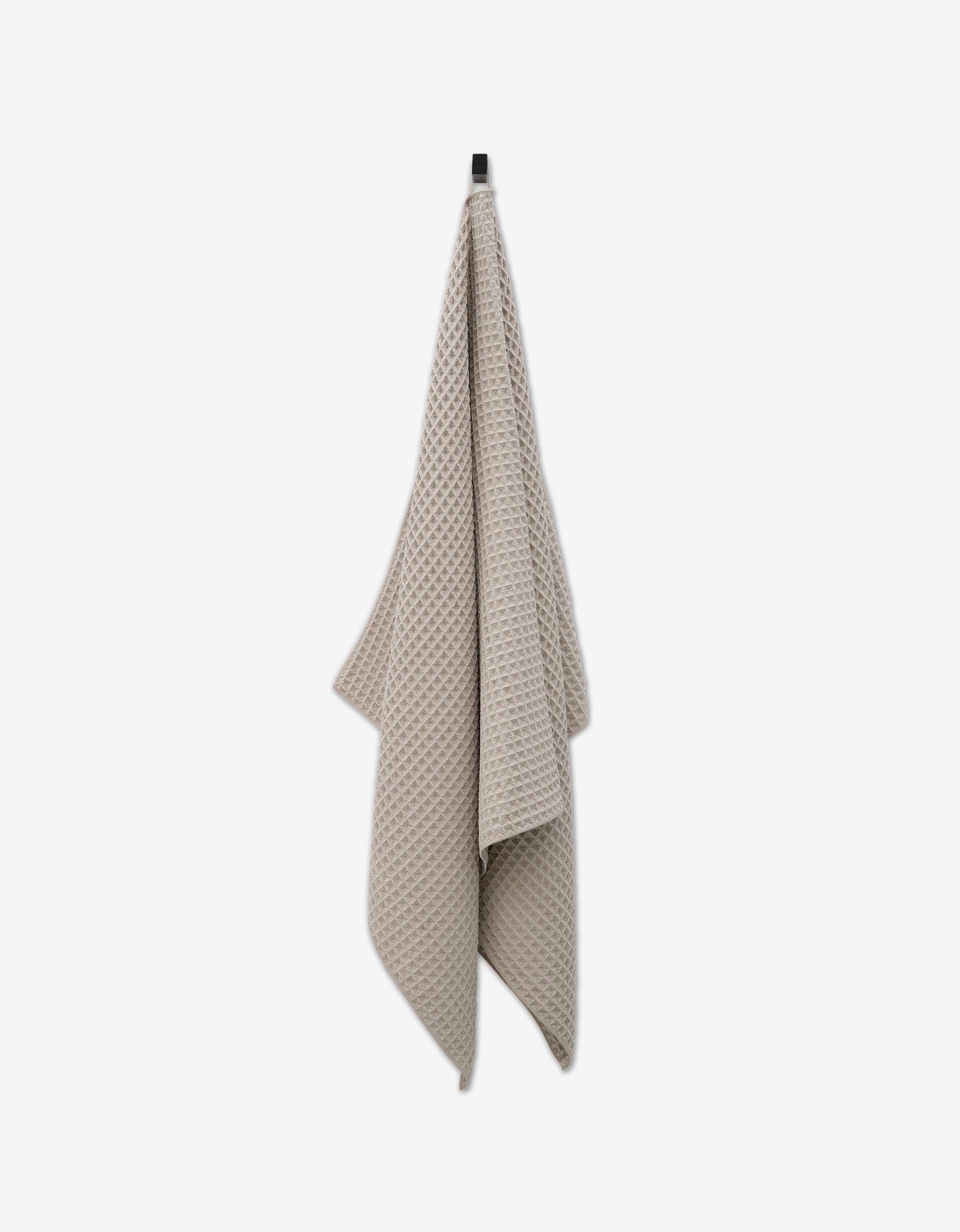 A taupe, waffle-textured GEOMETRY Waffle Bath Towel hangs from a black hook against a plain white background, combining elegance with daily comfort.