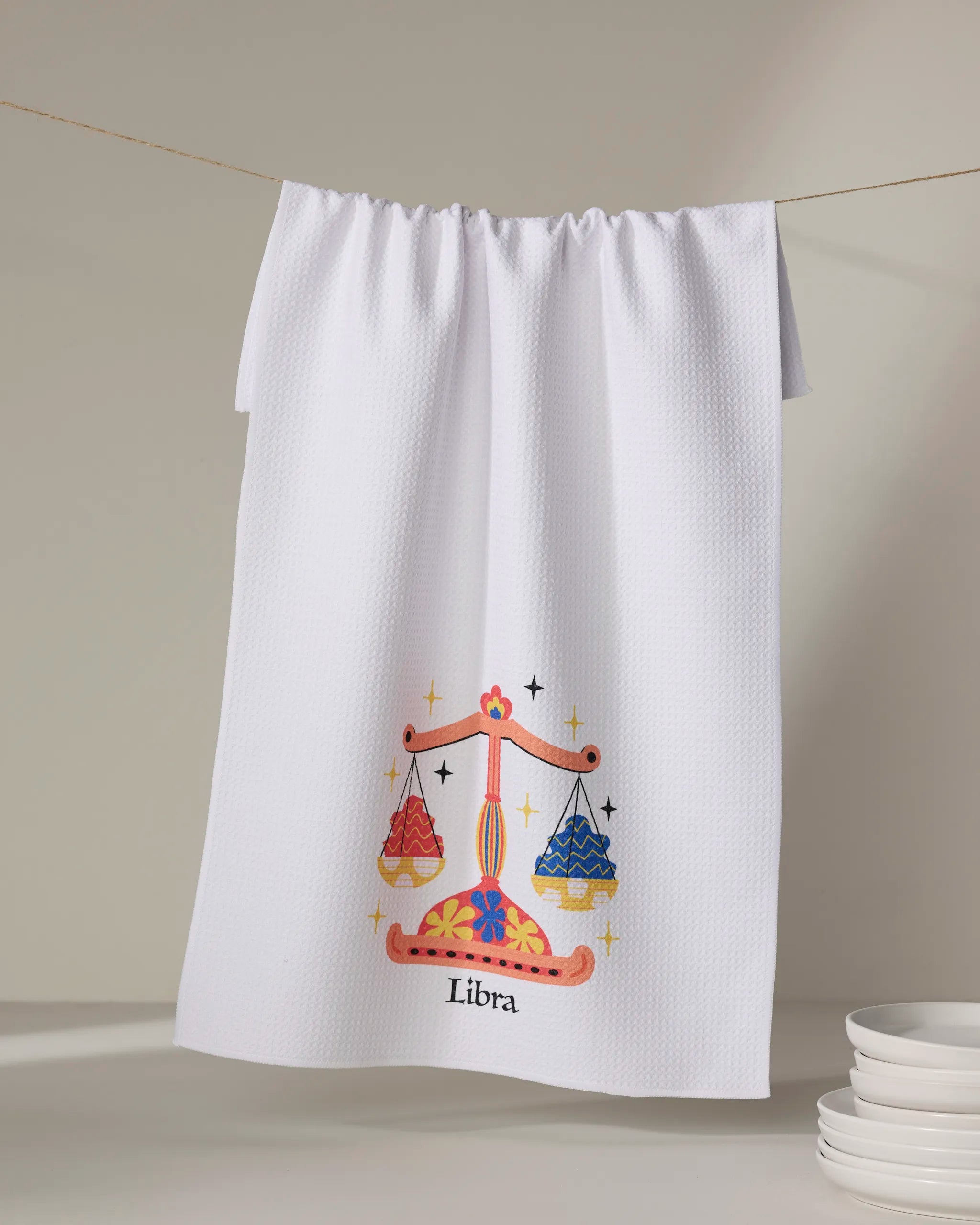A white recycled kitchen towel from Heather Zhou’s Charming Libra collection hangs on a line, showcasing a vibrant Libra illustration with a decorative scale and the word "Libra" below. Stacked white plates appear in the lower right corner.