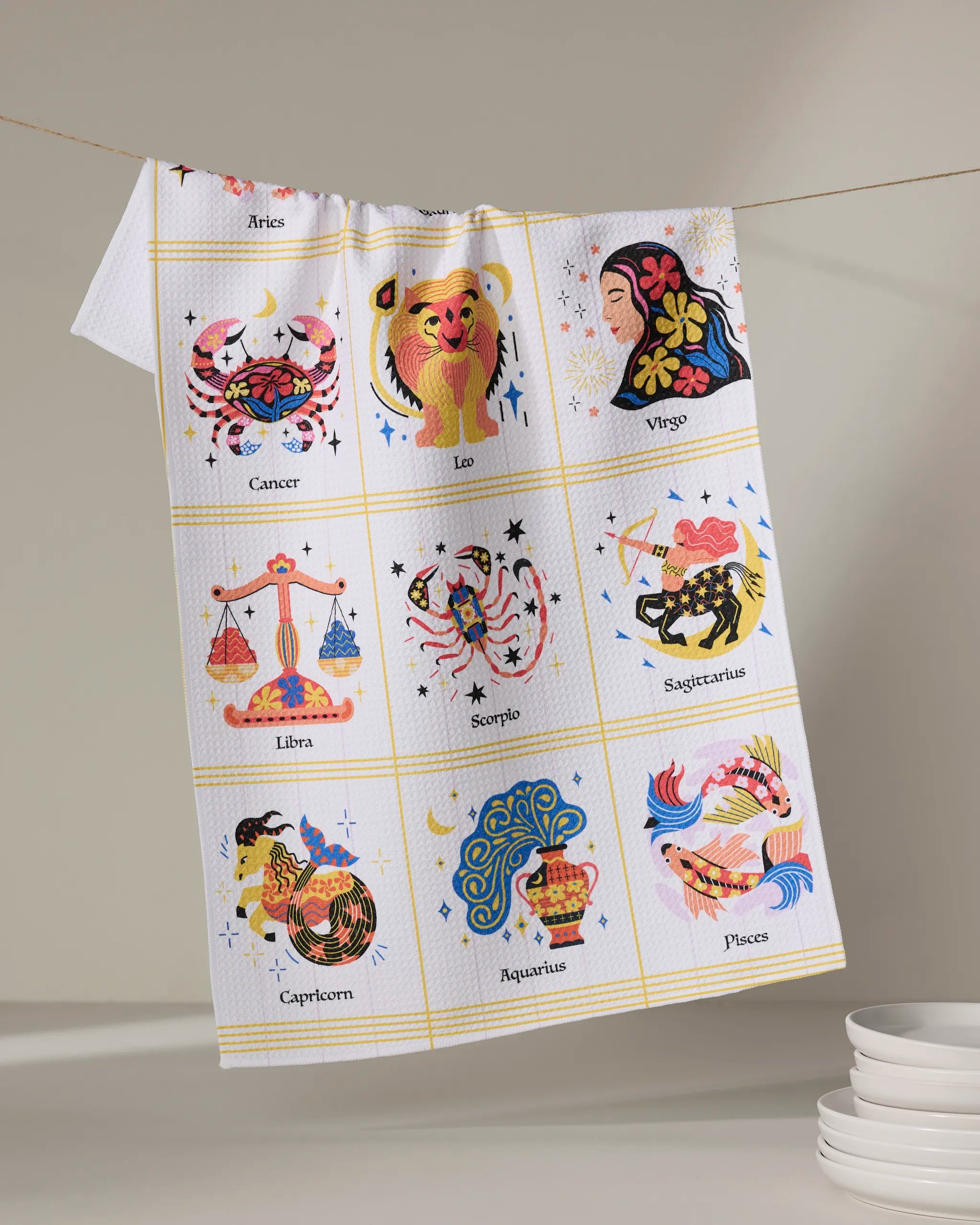 The Cosmic Harmony tea towel by Heather Zhou, made from recycled materials, features a vibrant 3x4 grid of labeled zodiac signs. Quick-drying and eco-friendly, it hangs beside stacked white plates.