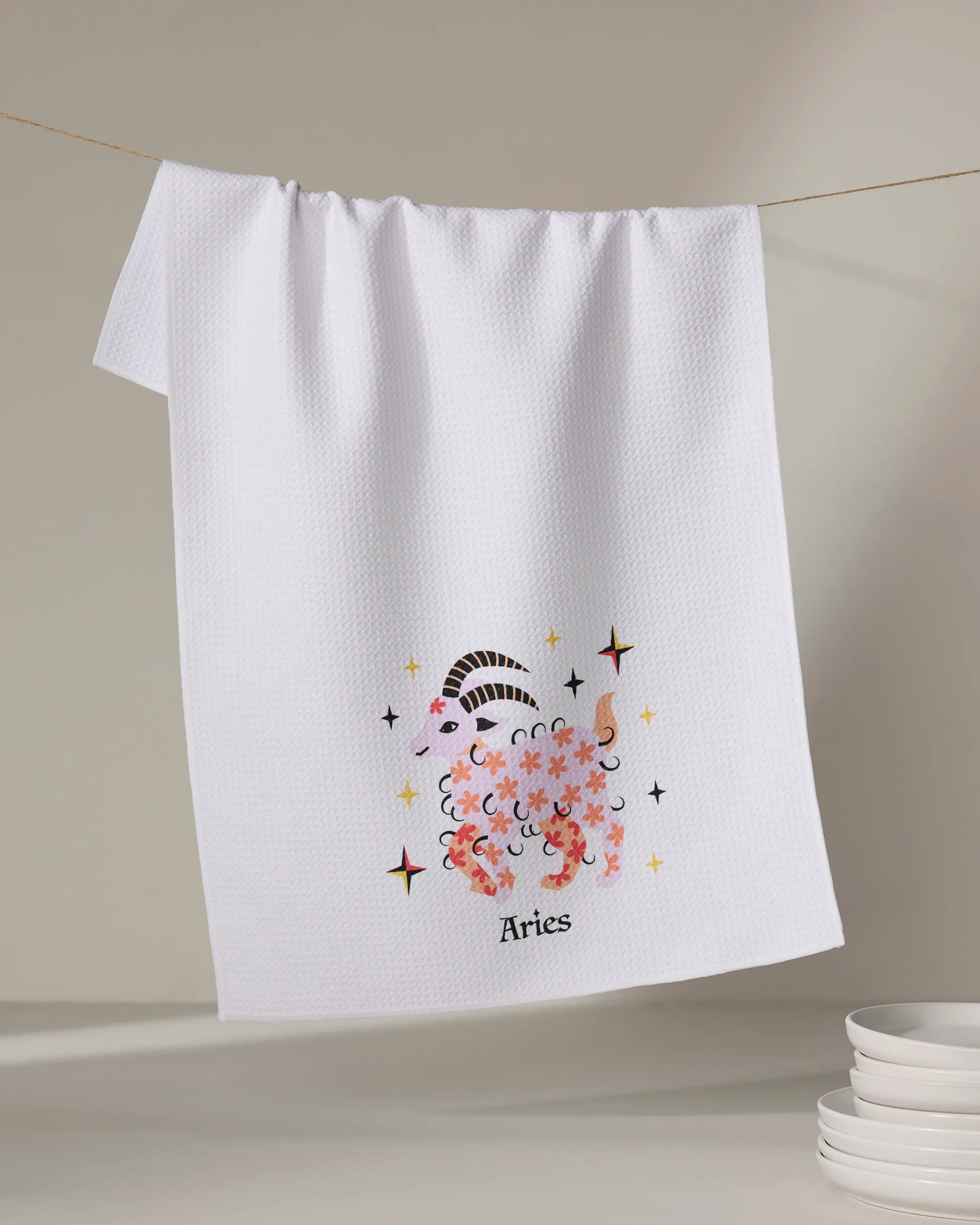 A white tea towel from Heather Zhou, called "Daring Aries," hangs on a line with a cute pink ram illustration, stars, and "Aries" beneath. A stack of white dishes sits in the lower right corner.
