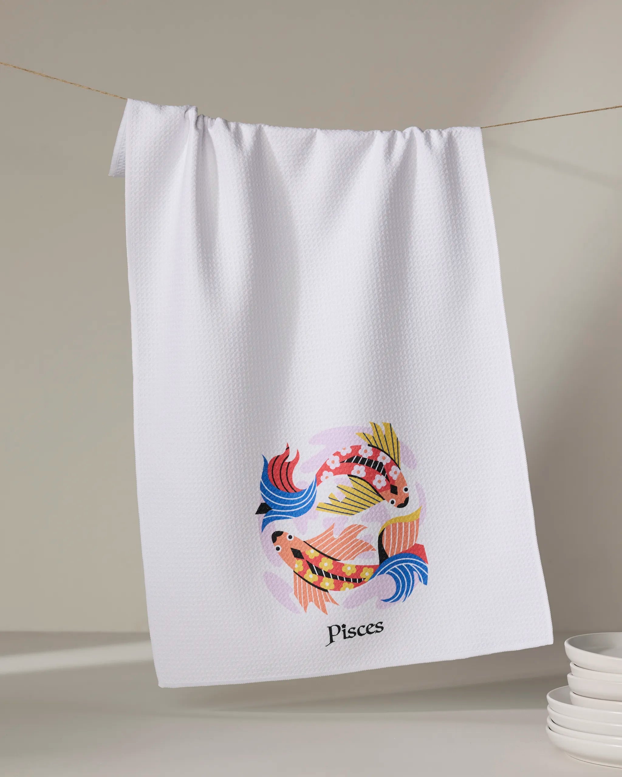 The Heather Zhou "Dreamy Pisces" tea towel, made from recycled materials, hangs on a line. Its colorful koi fish design brightens your kitchen, while ultra-absorbent fabric makes it perfect for drying dishes. White bowls are stacked nearby.