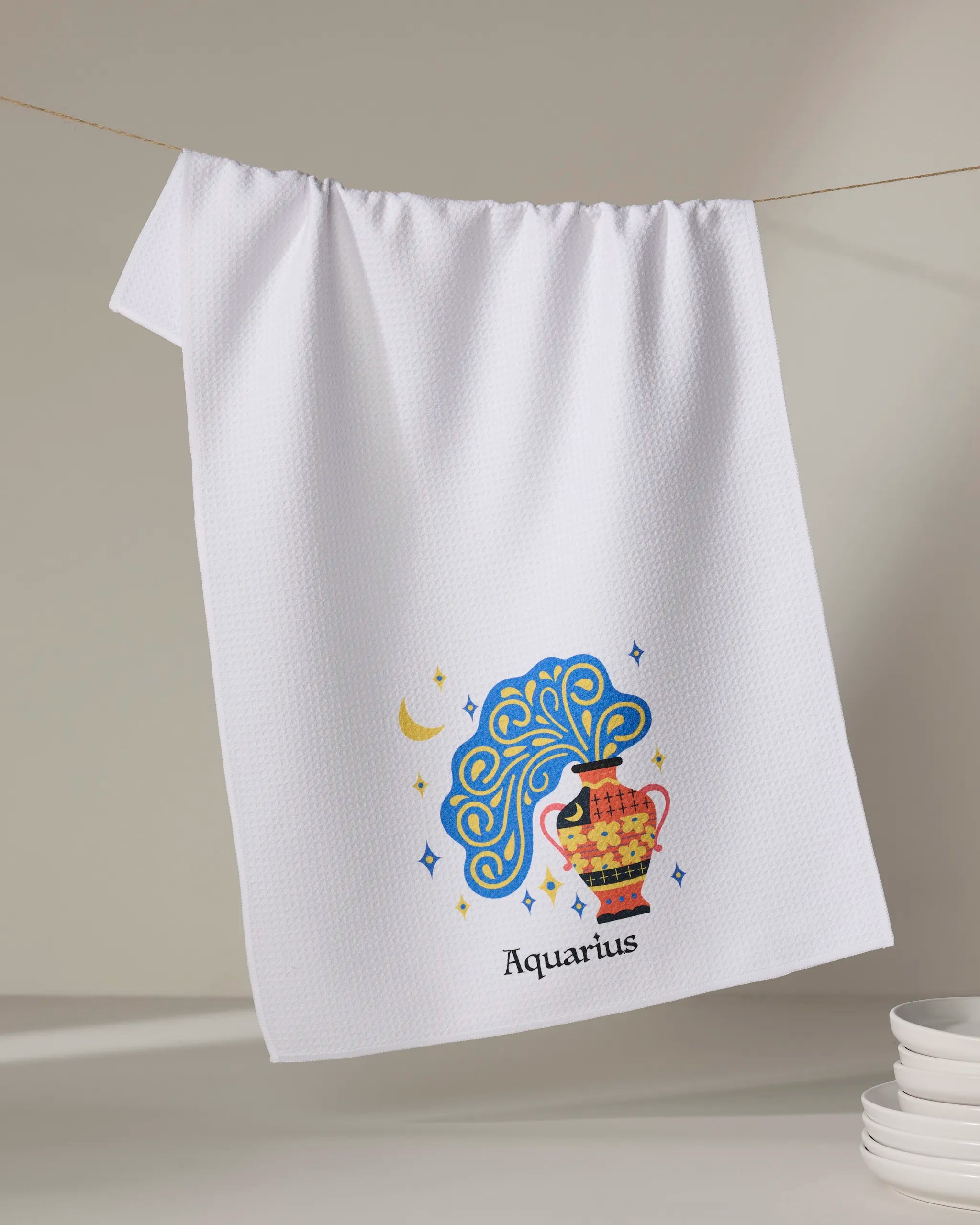 A white ultra-absorbent tea towel from Heather Zhou, "Free Aquarius," hangs on a line. It features a colorful illustration of a woman with a jug, star and moon motifs, and "Aquarius" text. Stacked plates appear in the corner.