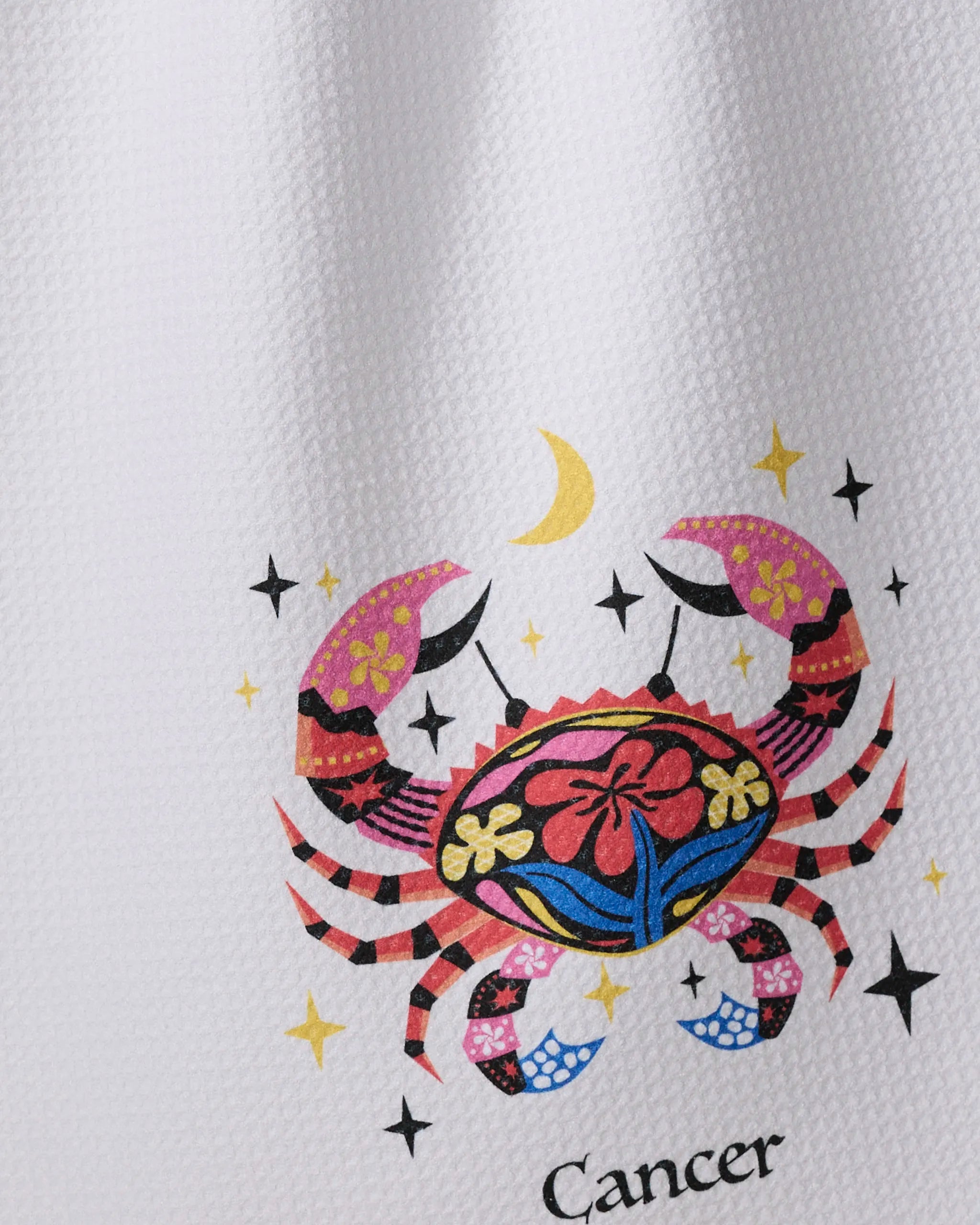 A vibrant kitchen towel by Heather Zhou features the "Gentle Cancer" design: a crab adorned with flowers and patterns amid stars and a crescent moon, with “Cancer” written beneath on textured white fabric.