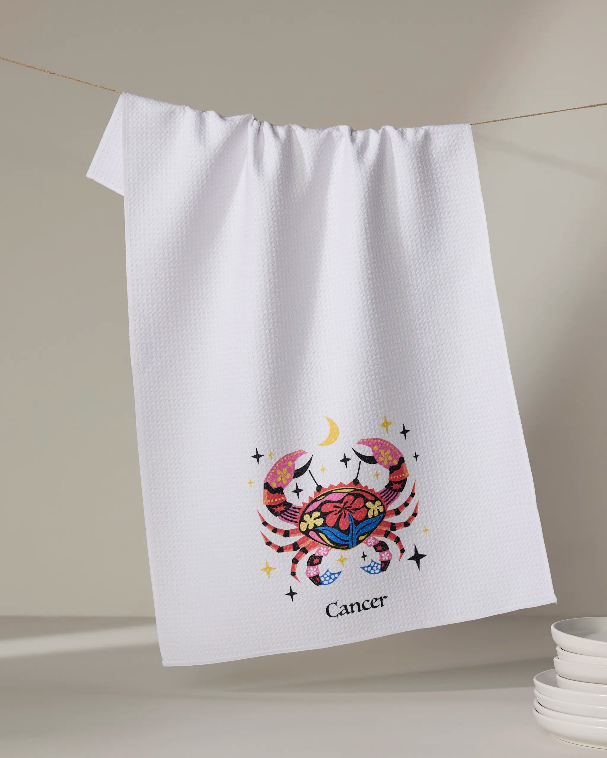 The Gentle Cancer towel by Heather Zhou hangs on a line, displaying a colorful crab with flowers, stars, and a crescent moon. "Cancer" is written below the design. White plates are stacked nearby.