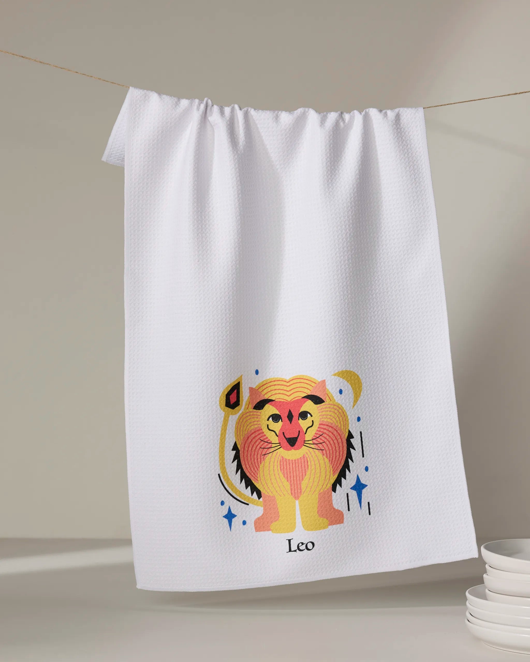 The Glowing Leo tea towel by Heather Zhou hangs on a line, displaying a vibrant cartoon lion and "Leo" below. Made from quick-drying recycled materials, it brightens your kitchen. A stack of white plates appears in the lower right corner.