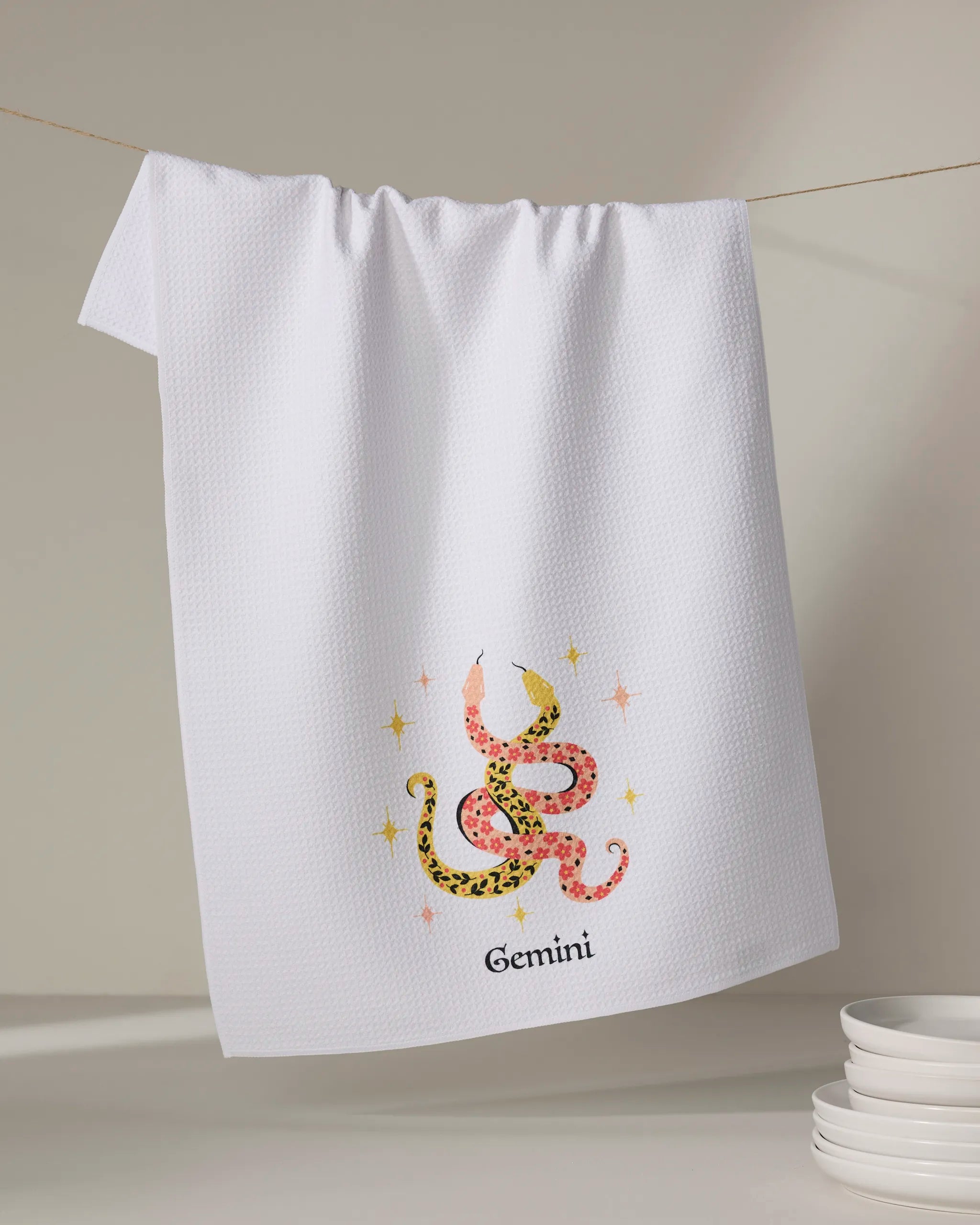 The Lucky Gemini tea towel by Heather Zhou hangs on a line, showcasing a pink and yellow patterned snake with stars and "Gemini" below. Ultra-absorbent and made from recycled materials, white plates are visible in the lower right corner.