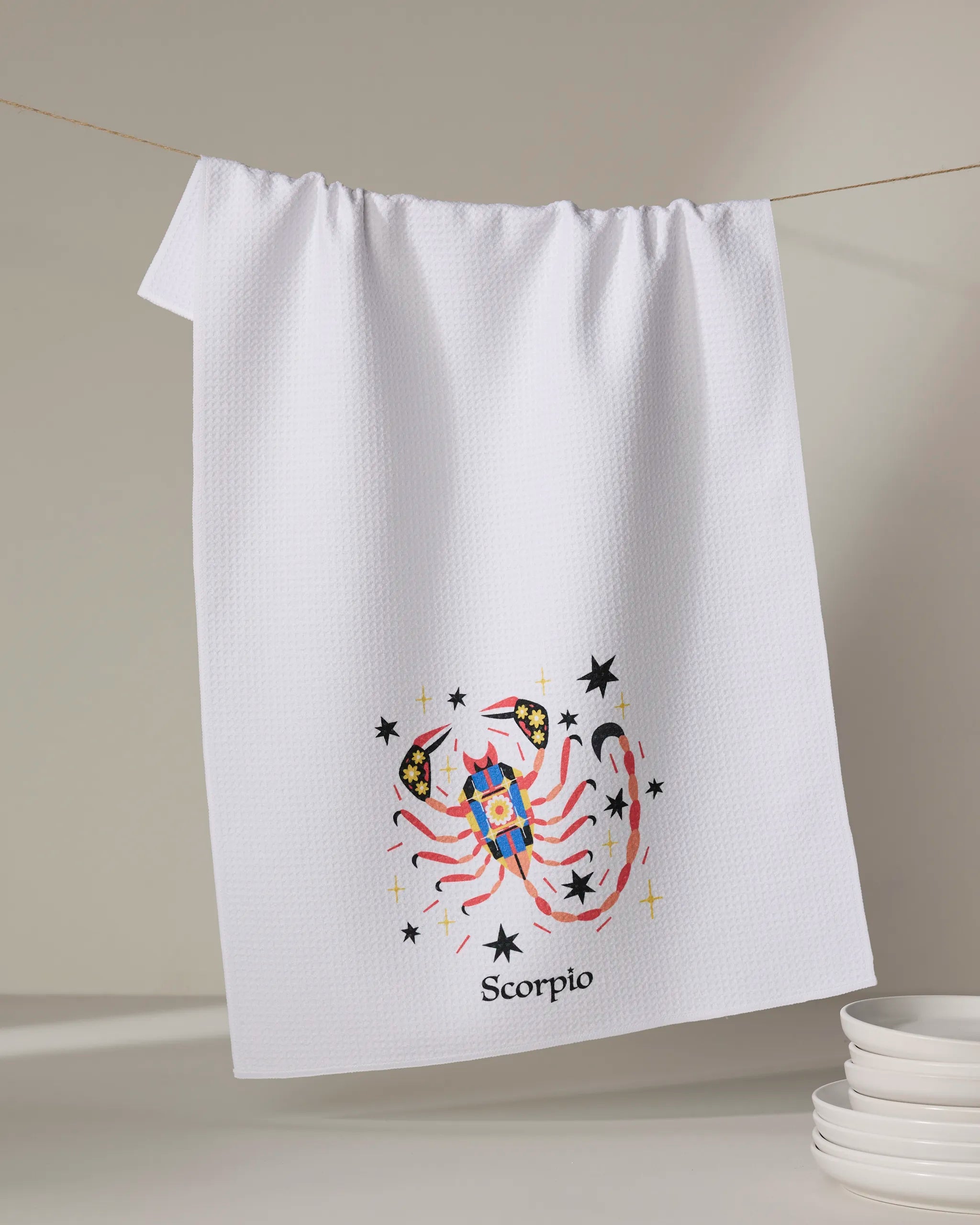 The "Mysterious Scorpio" tea towel by Heather Zhou, made from recycled materials, hangs on a line with a vibrant embroidered scorpion, stars, and “Scorpio” at the bottom. A stack of white dishes sits in the lower right corner.
