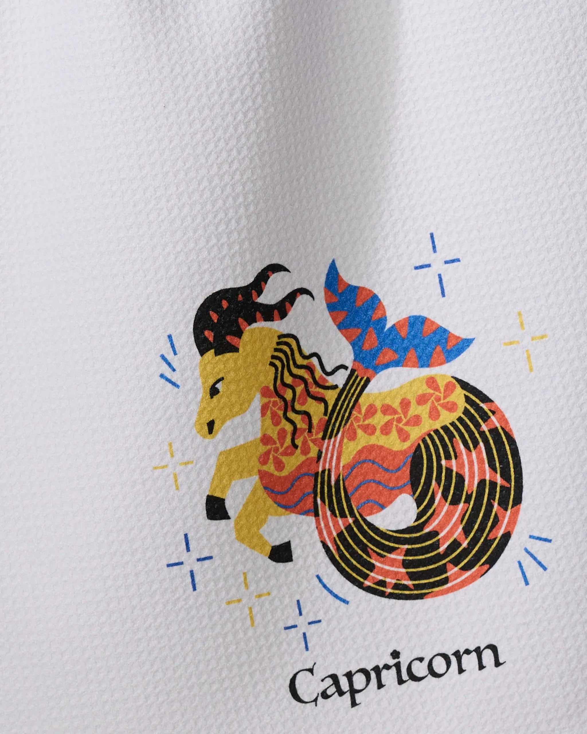 Steadfast Capricorn by Heather Zhou: a stylized goat with a fish tail and colorful patterns on a white textured background, with "Capricorn" below—perfect for a quick-drying tea towel.
