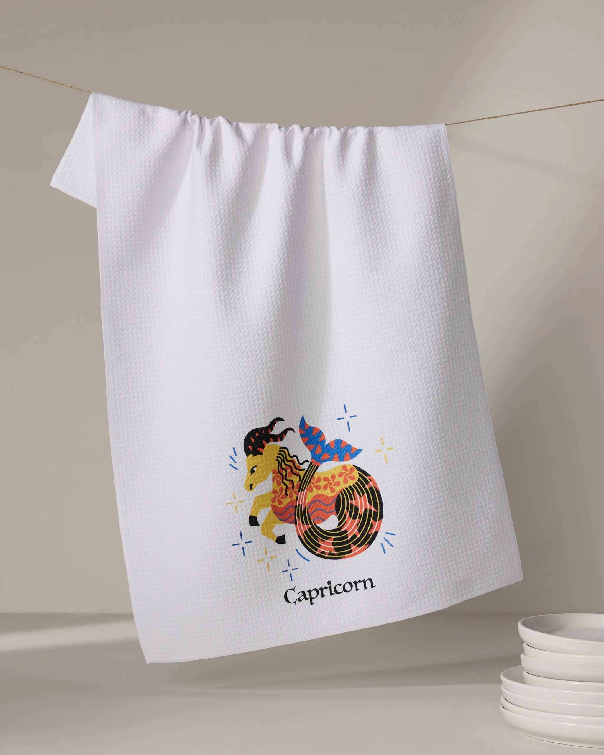 The Steadfast Capricorn tea towel by Heather Zhou hangs on a line, displaying a vibrant goat-fish illustration with “Capricorn” below. Made from quick-drying recycled material, white plates are stacked in the bottom right corner.
