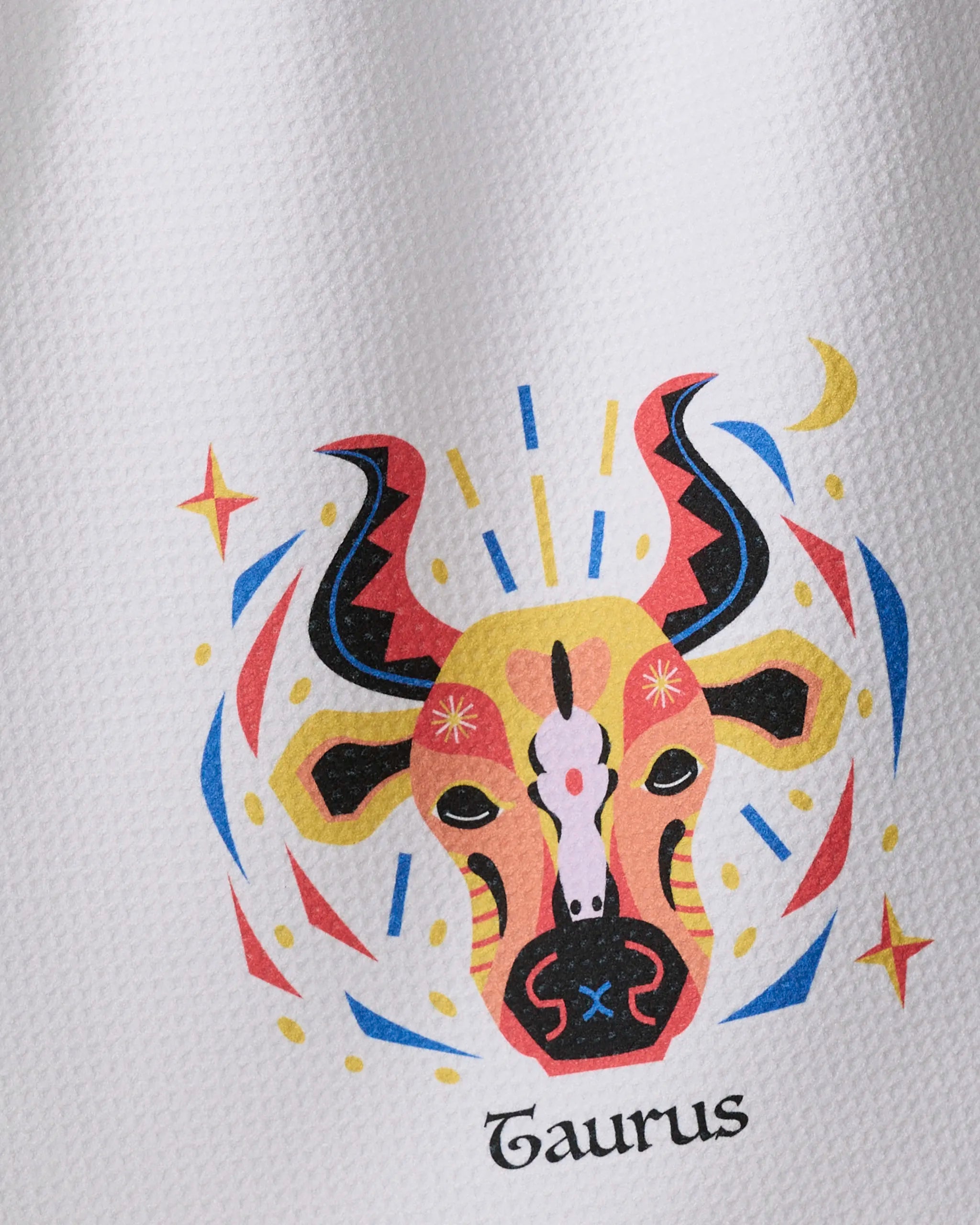 Heather Zhou’s Steady Taurus tea towel features a colorful bull’s head with abstract patterns, stars, and swirls on textured white fabric. “Taurus” is written below the vibrant illustration.