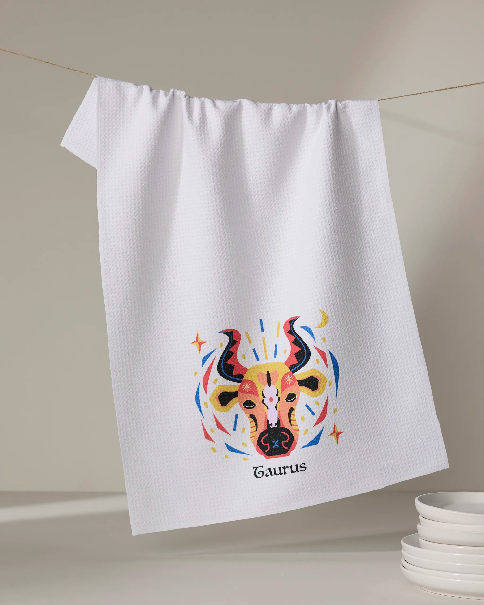 A white textured tea towel by Heather Zhou, named Steady Taurus, hangs on a line showcasing a vibrant Taurus bull illustration. "Taurus" is printed below the design, and white plates are stacked in the bottom right corner.