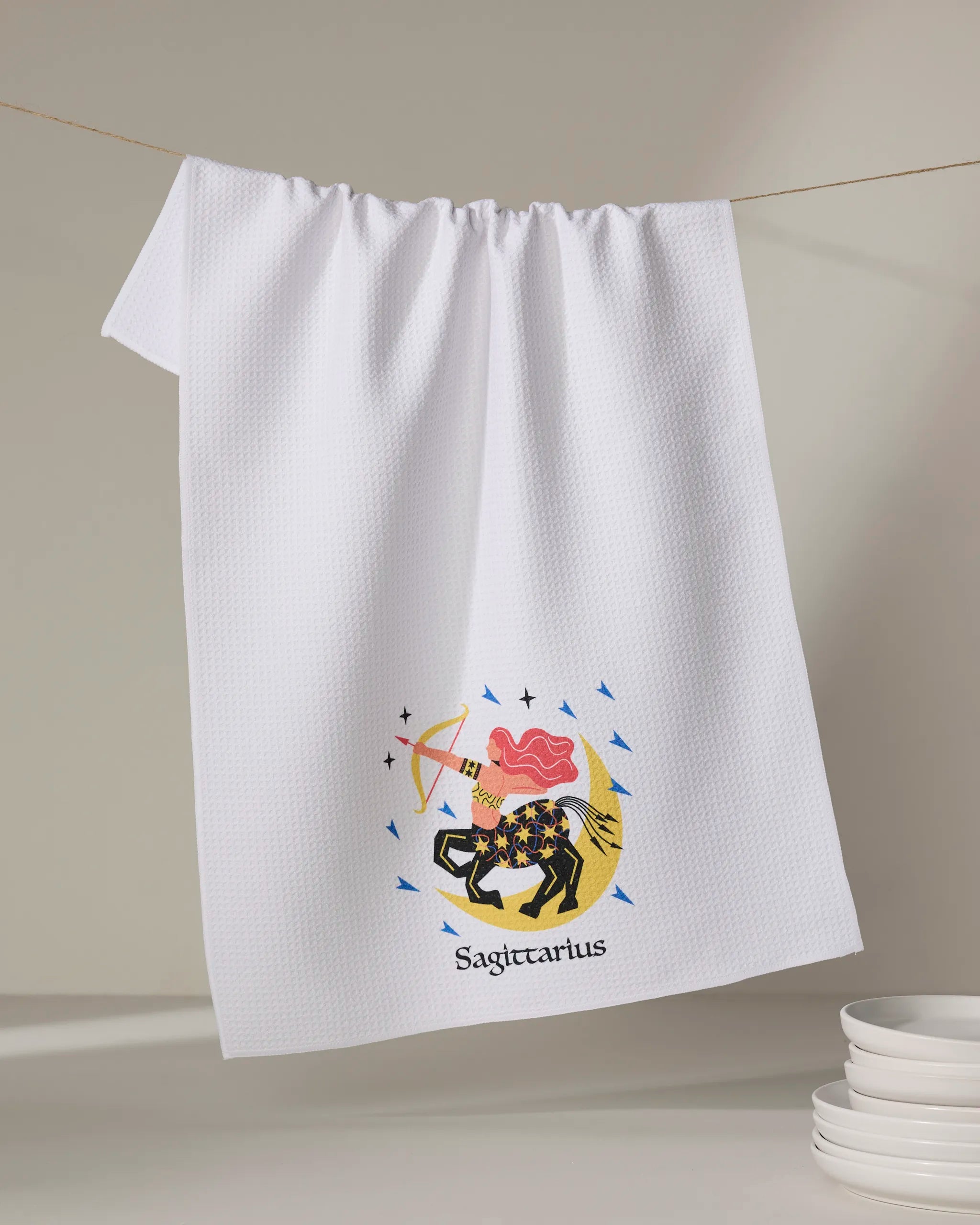 The Wild Sagittarius tea towel by Heather Zhou, made from recycled white fabric, hangs on a line featuring a colorful centaur with bow and arrow, pink cloud and stars, and "Sagittarius" below. Stacked dishes are nearby.