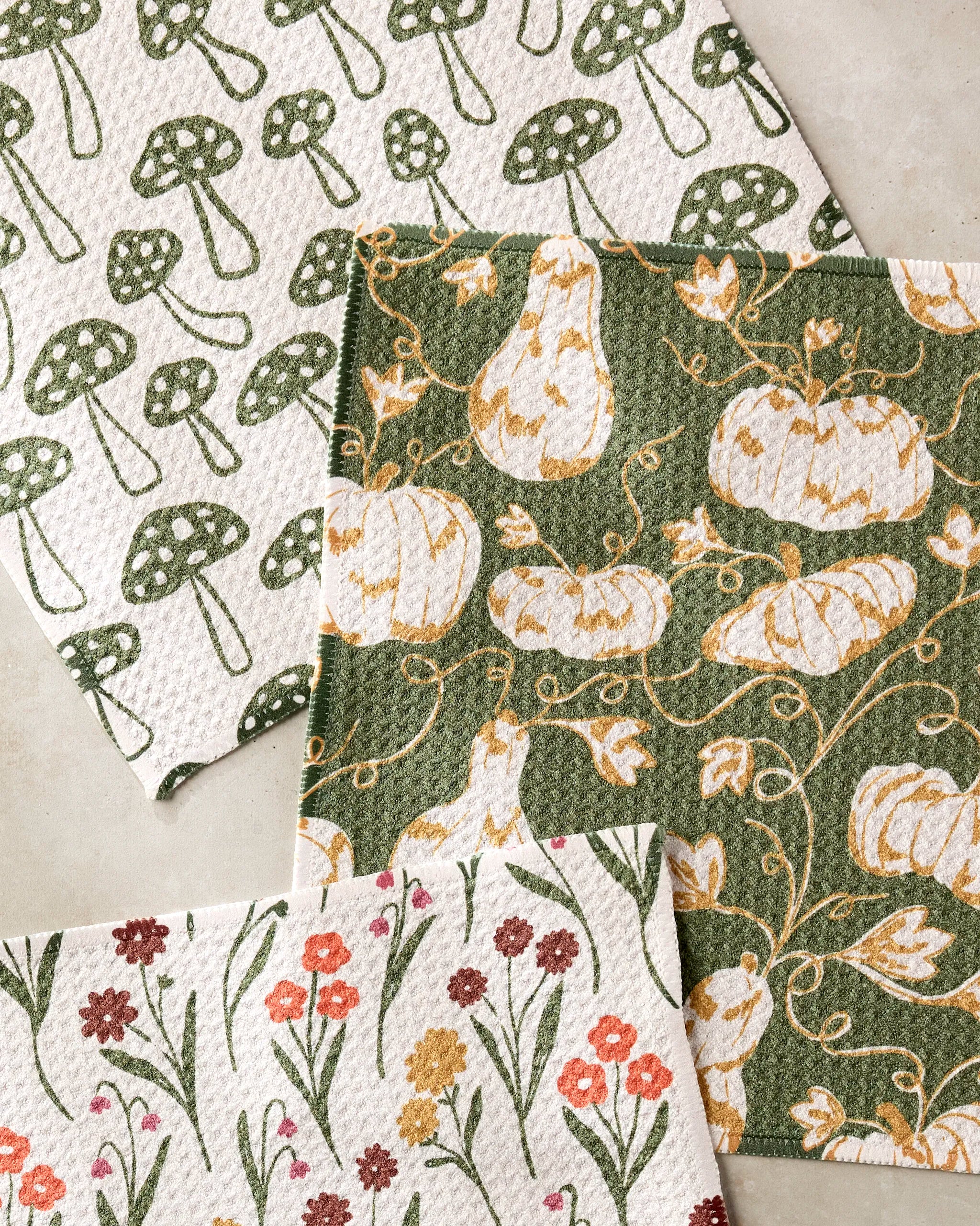 The Kayla Ann Woodland Trail set includes three stylish cloths: a green mushroom super-absorbent dishtowel, one with pumpkins and vines on dark green, and one with red, pink, and orange flowers on cream—perfect for any kitchen.