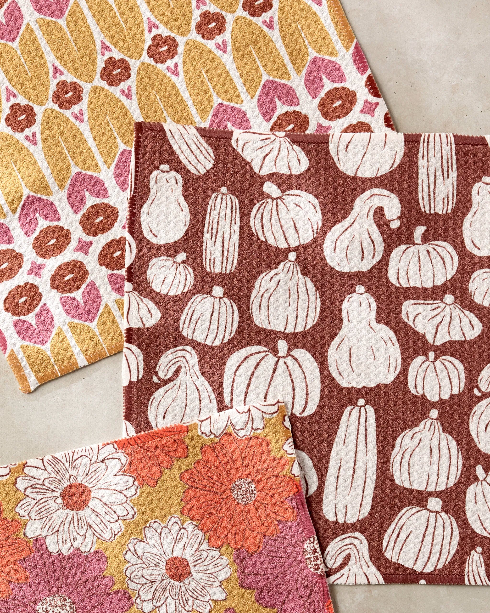 Three Zinnia Forever cloths by Kayla Ann rest on a neutral surface: one with yellow and pink abstract shapes, one featuring white gourds on rust, and another with daisies in yellow, white, and coral—all crafted from recycled materials.