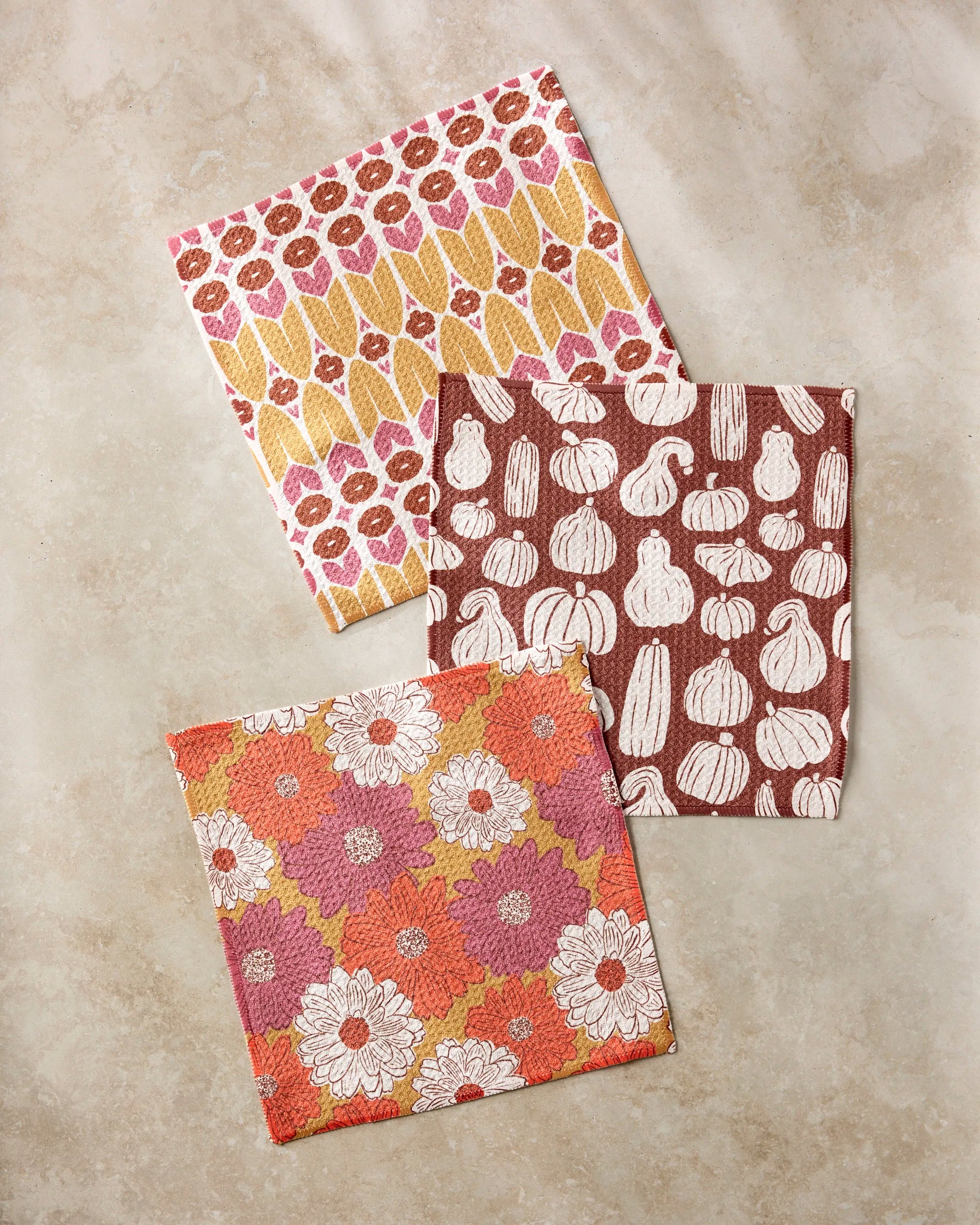 The Zinnia Forever dishcloth set by Kayla Ann features three vibrant cloths—yellow and pink corn, white gourds on brown, and floral designs—each crafted from recycled materials for a colorful, eco-friendly touch.