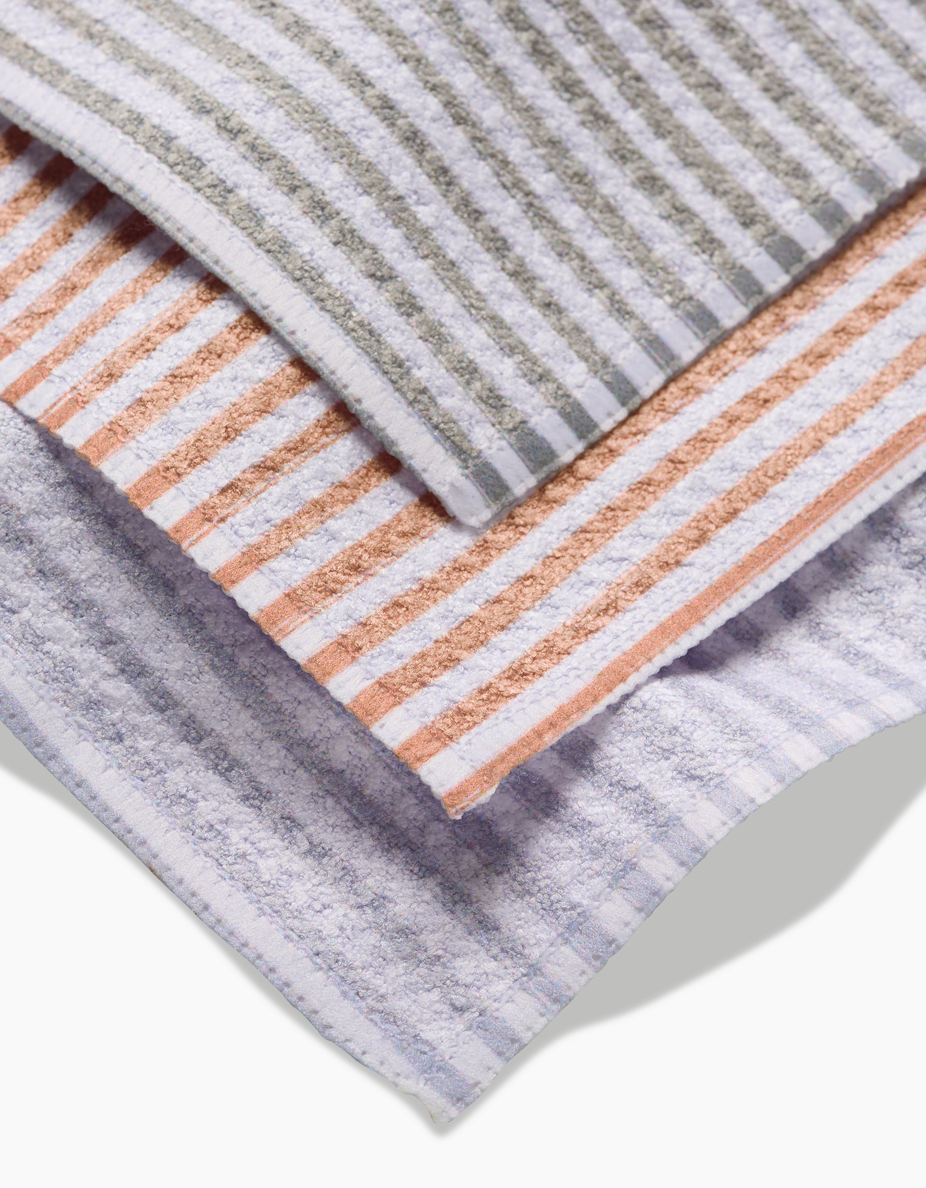 Three À La Carte towels by Kailee Wright, made from recycled materials, are stacked: the top has gray and white stripes, the middle is orange and white, and the bottom features light purple and white stripes.