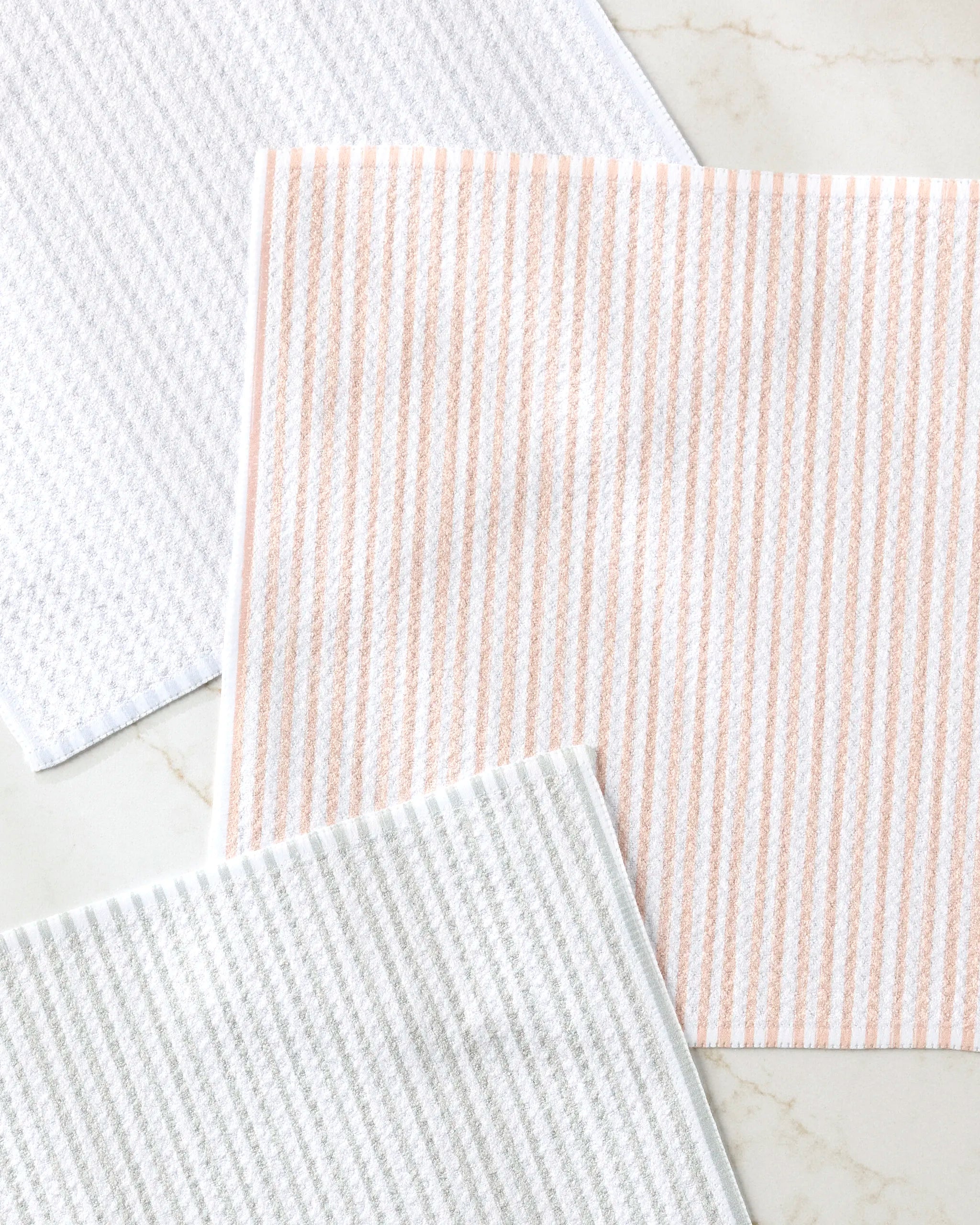 The À La Carte dishcloth set by Kailee Wright includes three rectangular cloths made from recycled materials—one white, one with peach stripes, and one with light gray stripes—each featuring a stylish textured, striped pattern.
