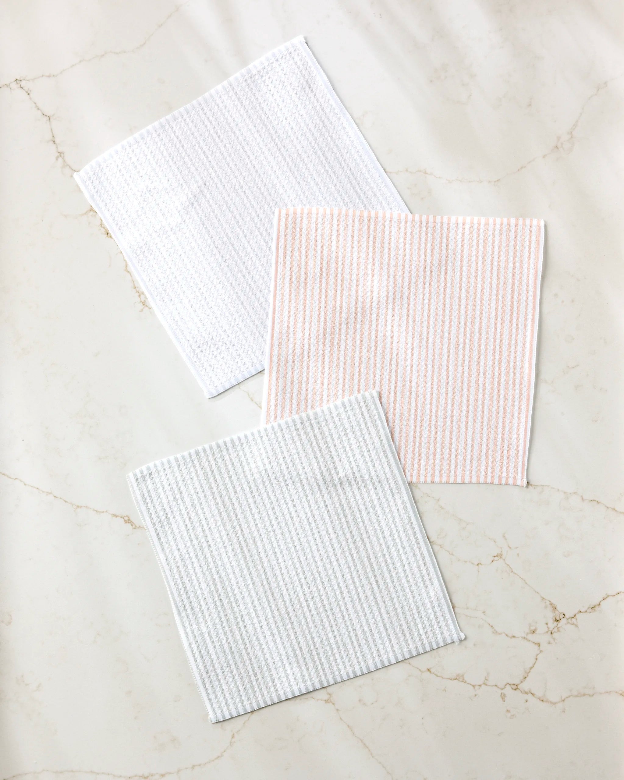 Three square À La Carte cloth napkins by Kailee Wright, made from recycled materials, are displayed on a light marble surface: one plain white, one with light pink stripes, and one with light gray stripes.
